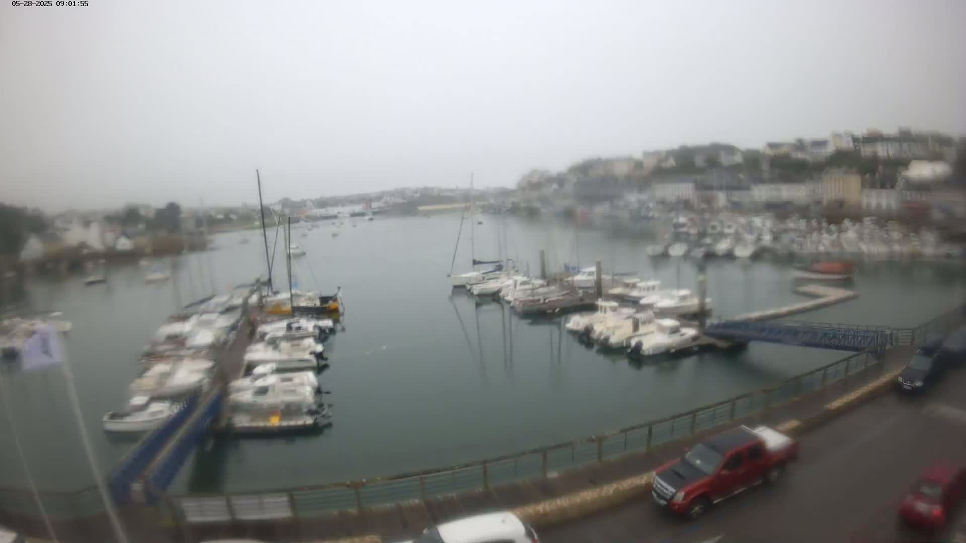 A harbor filled with numerous sailboats and motorboats is seen on an overcast day, with cars parked along the waterfront.