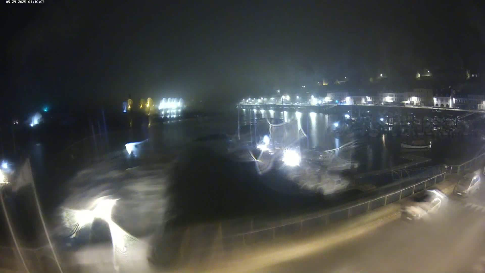 A nighttime view of a harbor filled with boats under foggy or misty conditions, with a town's lights visible in the background.