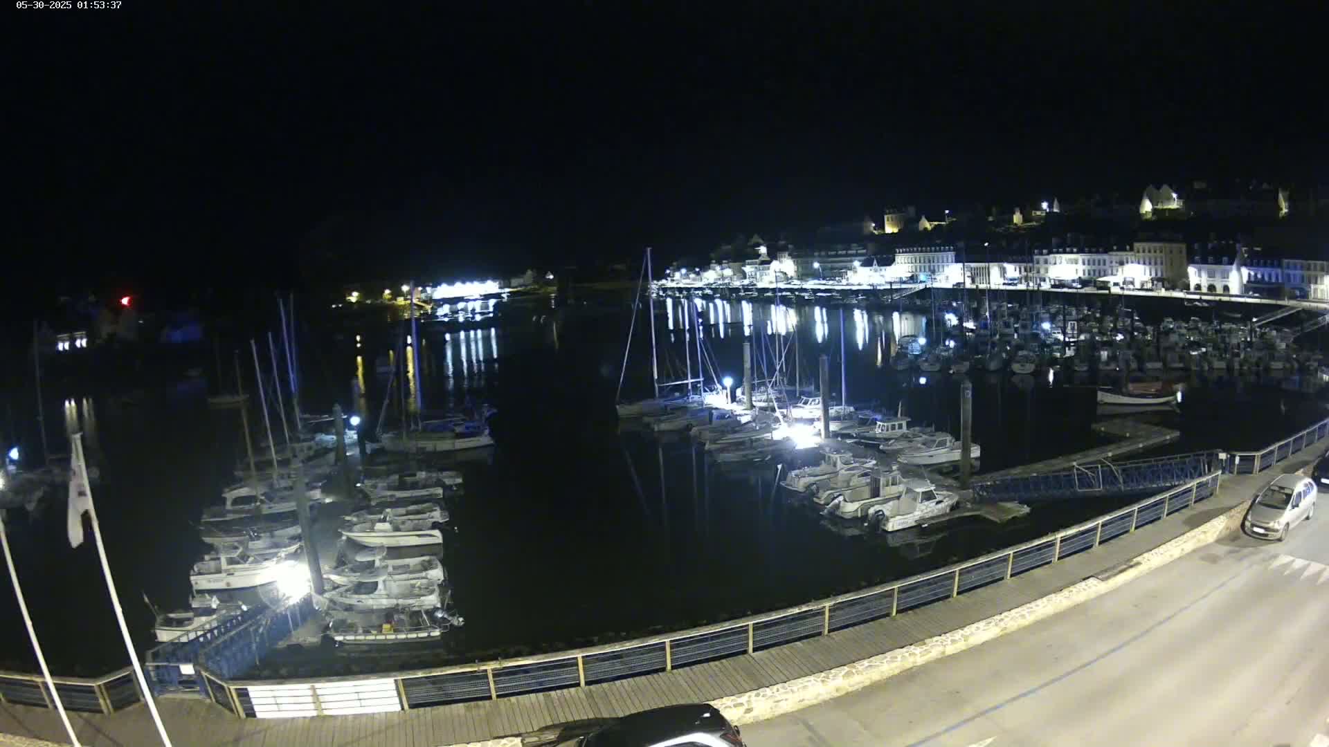 A nighttime view of a harbor filled with docked sailboats and motorboats, illuminated by lights from buildings on the opposite shore.