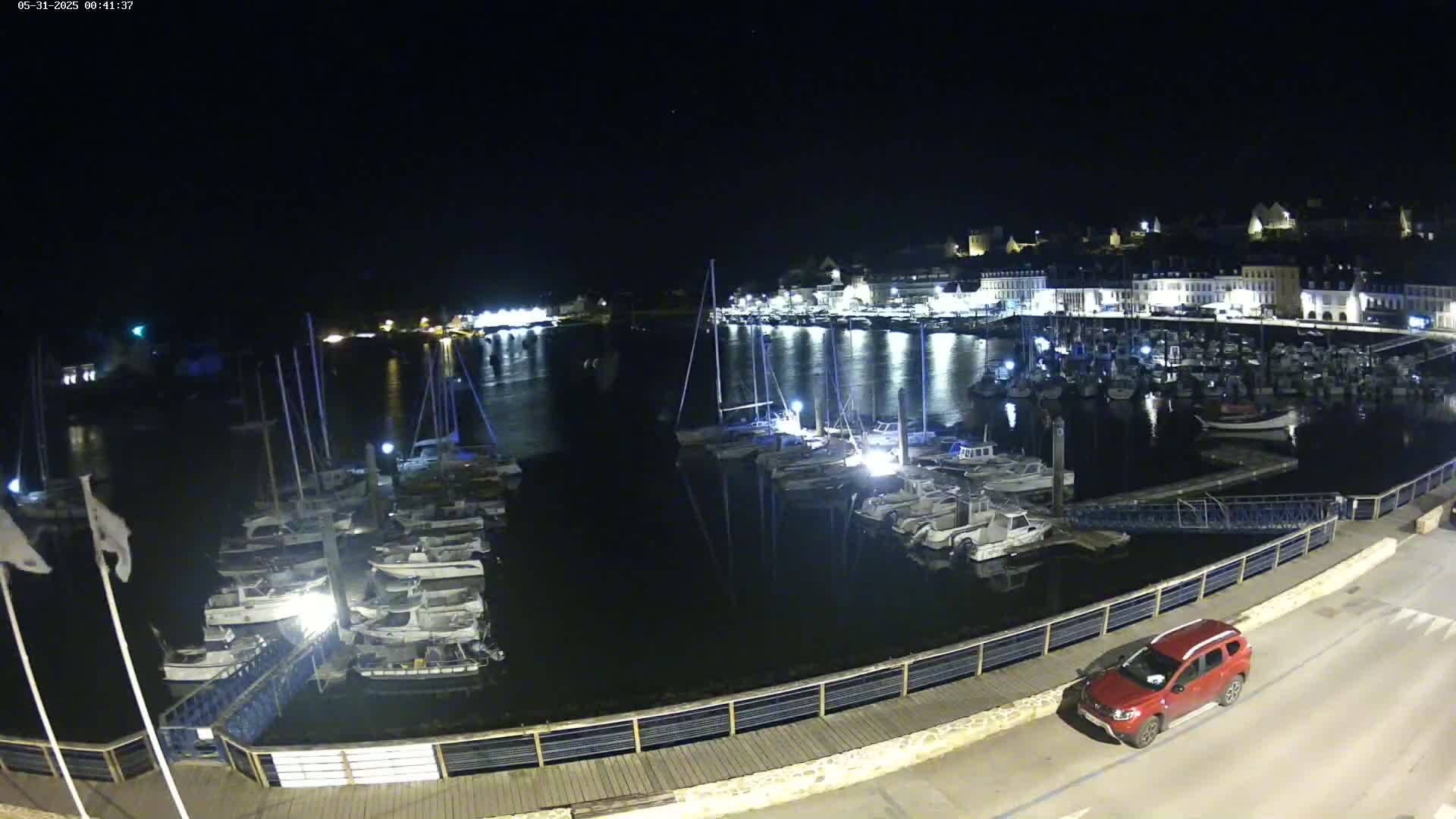 A nighttime view of a harbor filled with boats, illuminated by lights reflecting on the calm water, with a red car parked nearby on a paved area.