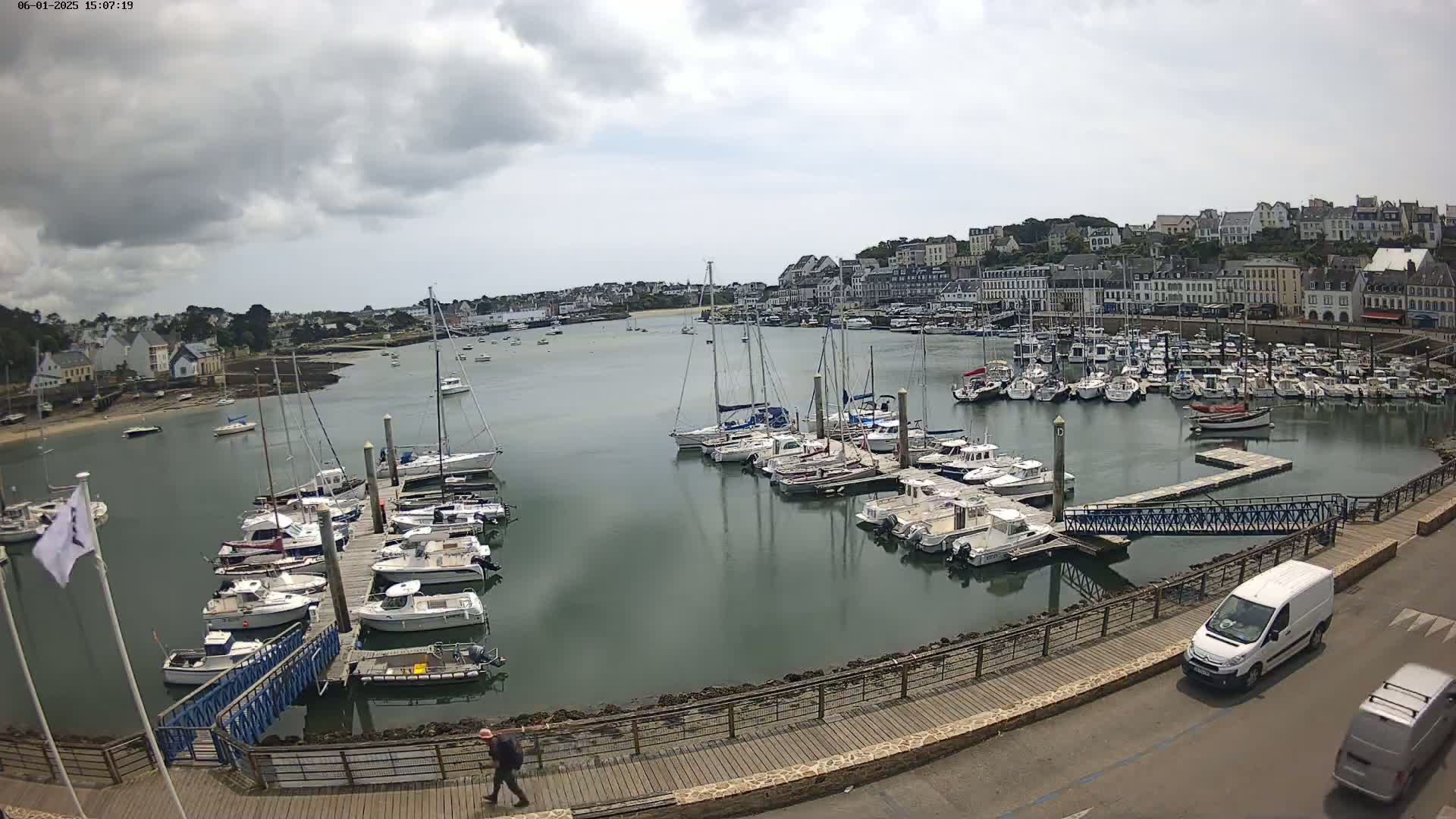 A harbor filled with numerous sailboats and motorboats under a partly cloudy sky, with a town and buildings visible in the background.