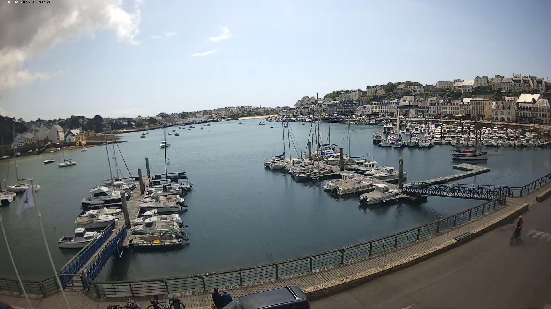 A harbor filled with numerous boats and yachts is seen on a partly sunny day, with a town's buildings visible in the background.