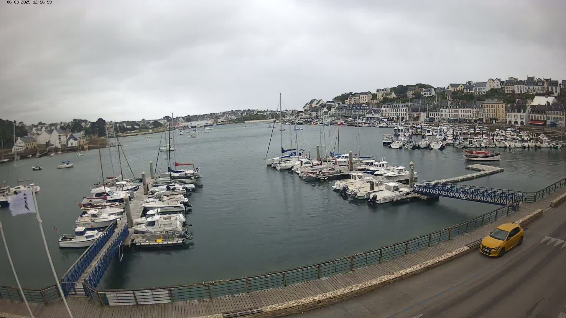 A harbor filled with numerous boats and yachts is shown under an overcast sky, with a yellow car parked on a nearby road.