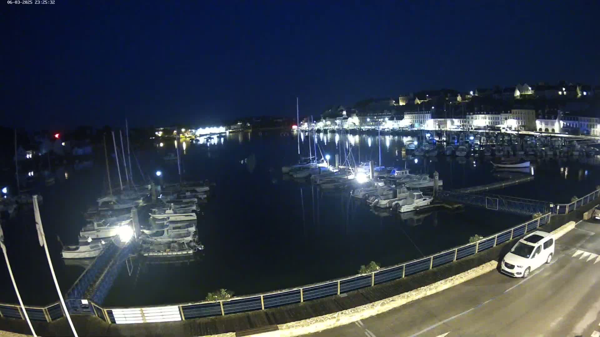 A nighttime view of a harbor filled with docked boats, illuminated by lights from the town on the opposite shore under a dark sky.