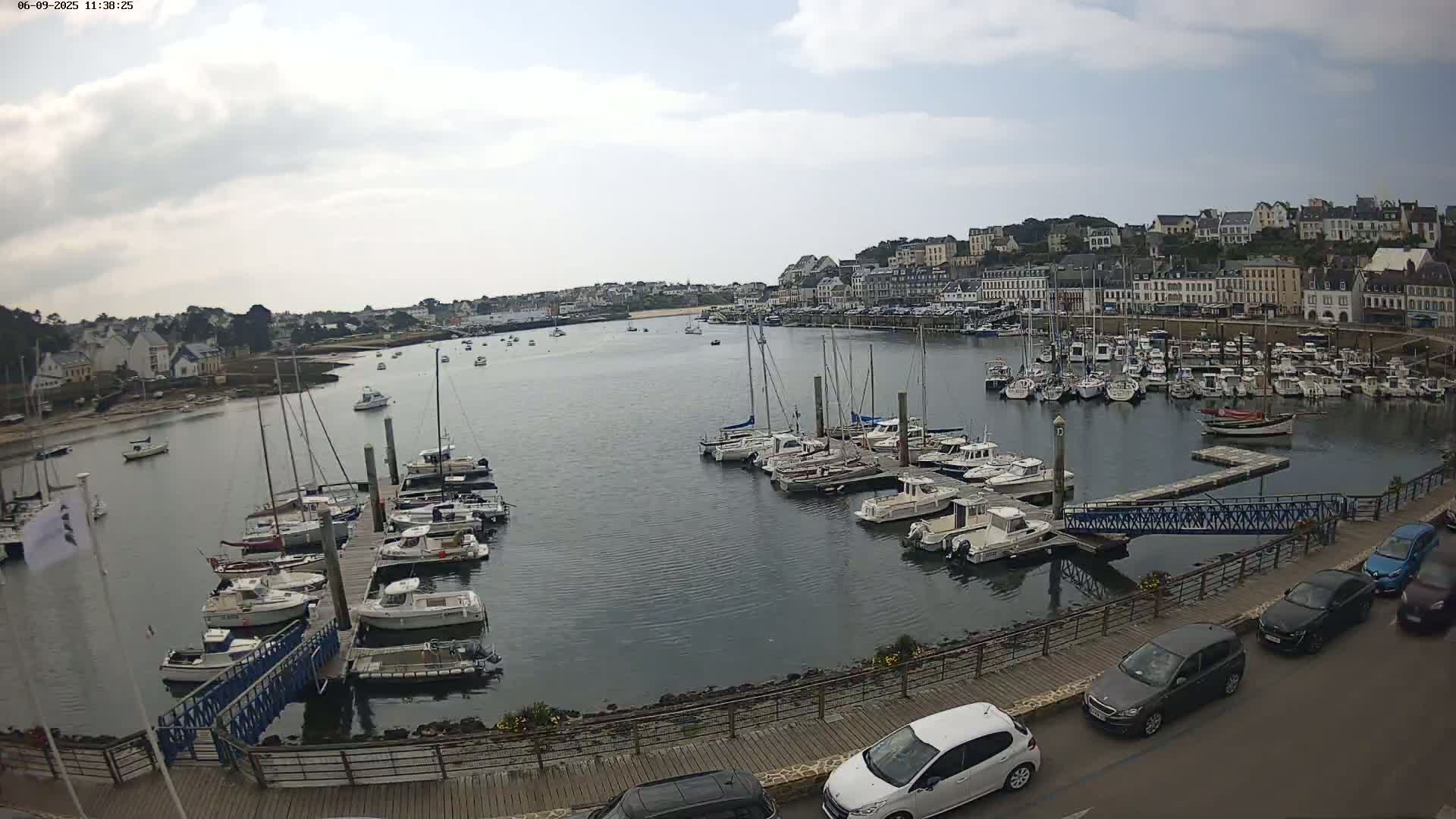 A harbor filled with numerous boats and yachts under a partly cloudy sky, with cars parked along a nearby roadway overlooking the water.