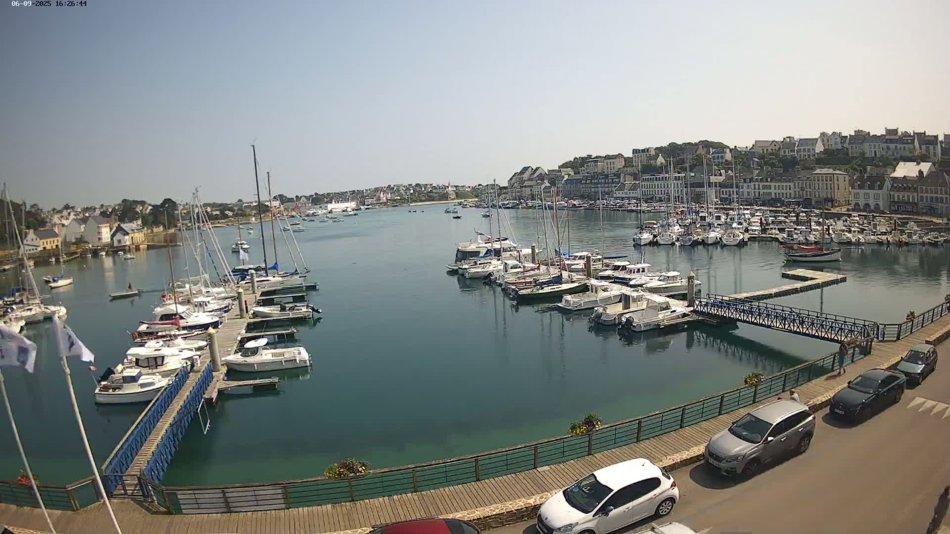 A harbor filled with numerous sailboats and motorboats on a sunny day, with a town in the background and cars parked along a waterfront walkway.