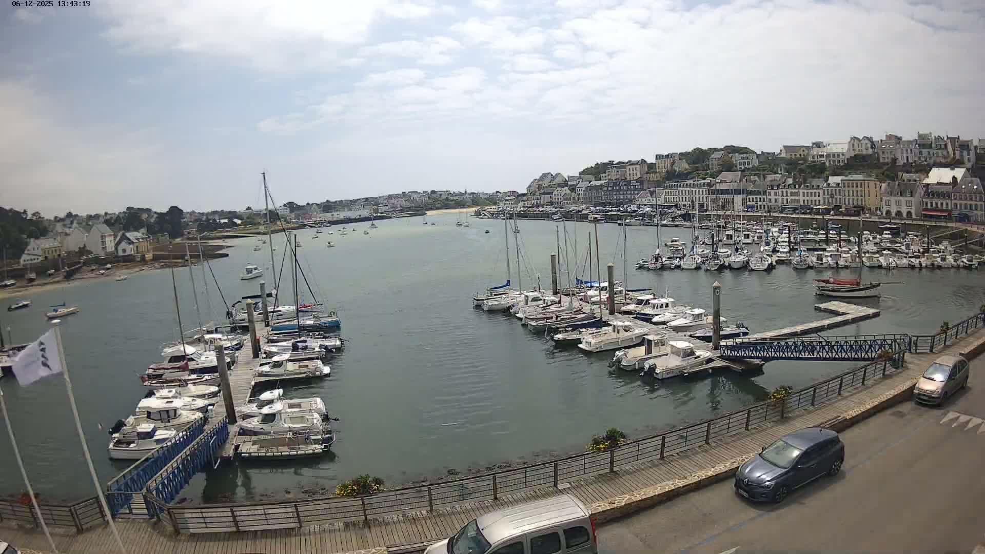 A harbor filled with numerous boats on a partly cloudy day, with cars parked along a nearby roadway and buildings lining the shore in the background.