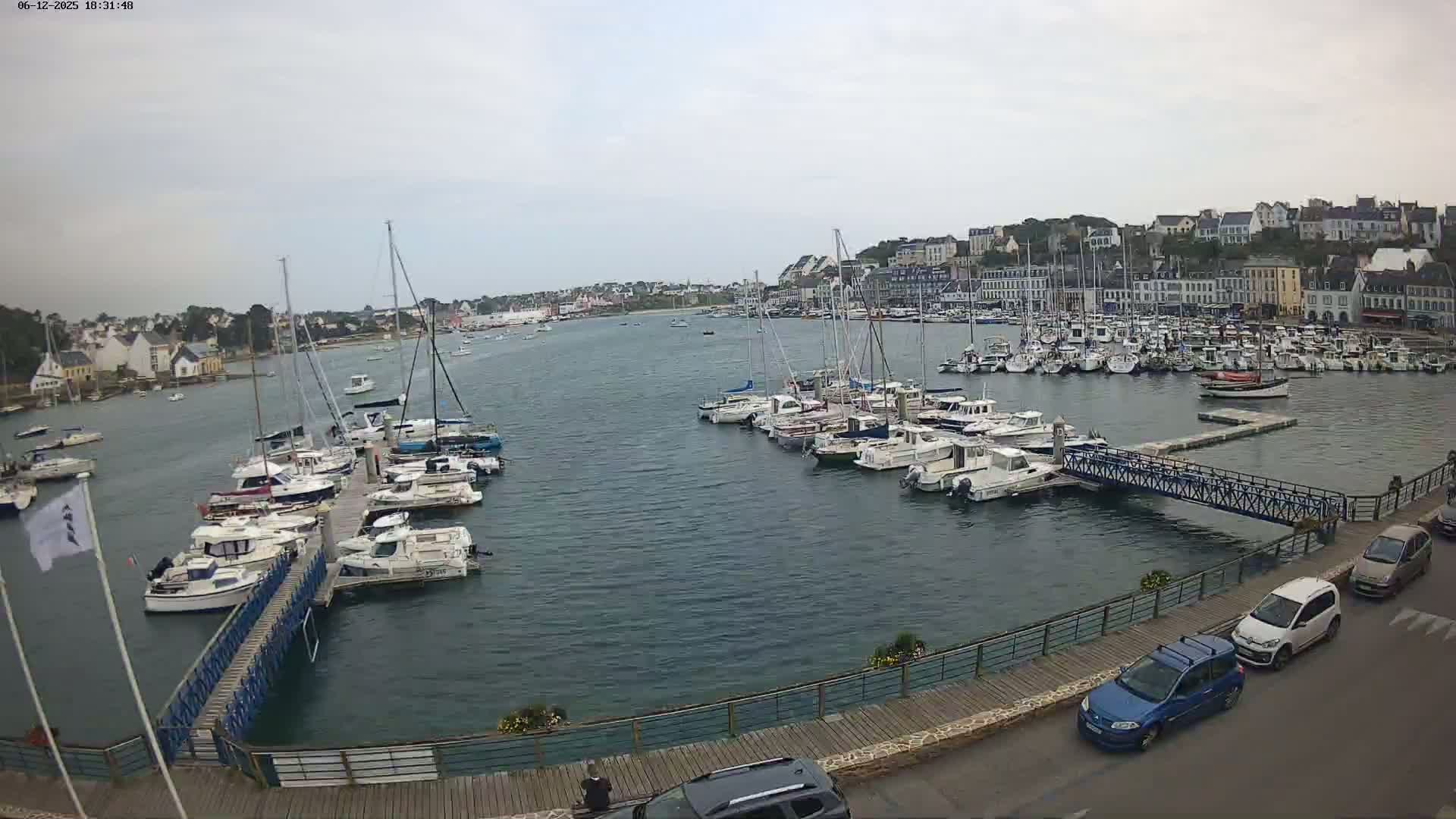 A harbor filled with numerous boats and yachts under a cloudy sky, with a town visible in the background and cars parked along a waterfront walkway.