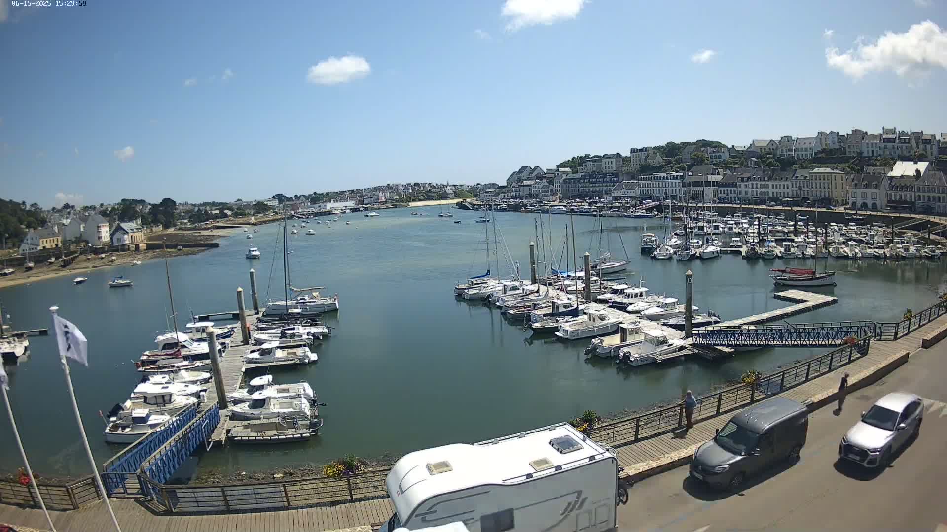A harbor filled with numerous boats and yachts under a mostly sunny sky, with a town and buildings visible in the background.