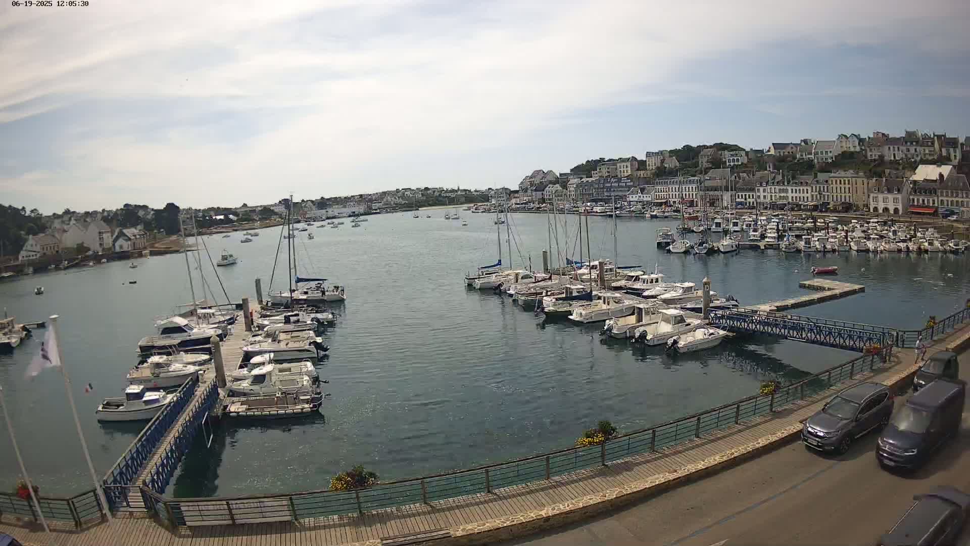 A harbor filled with numerous boats and yachts is seen on a partly cloudy day, with a town of white buildings situated on a hill in the background.