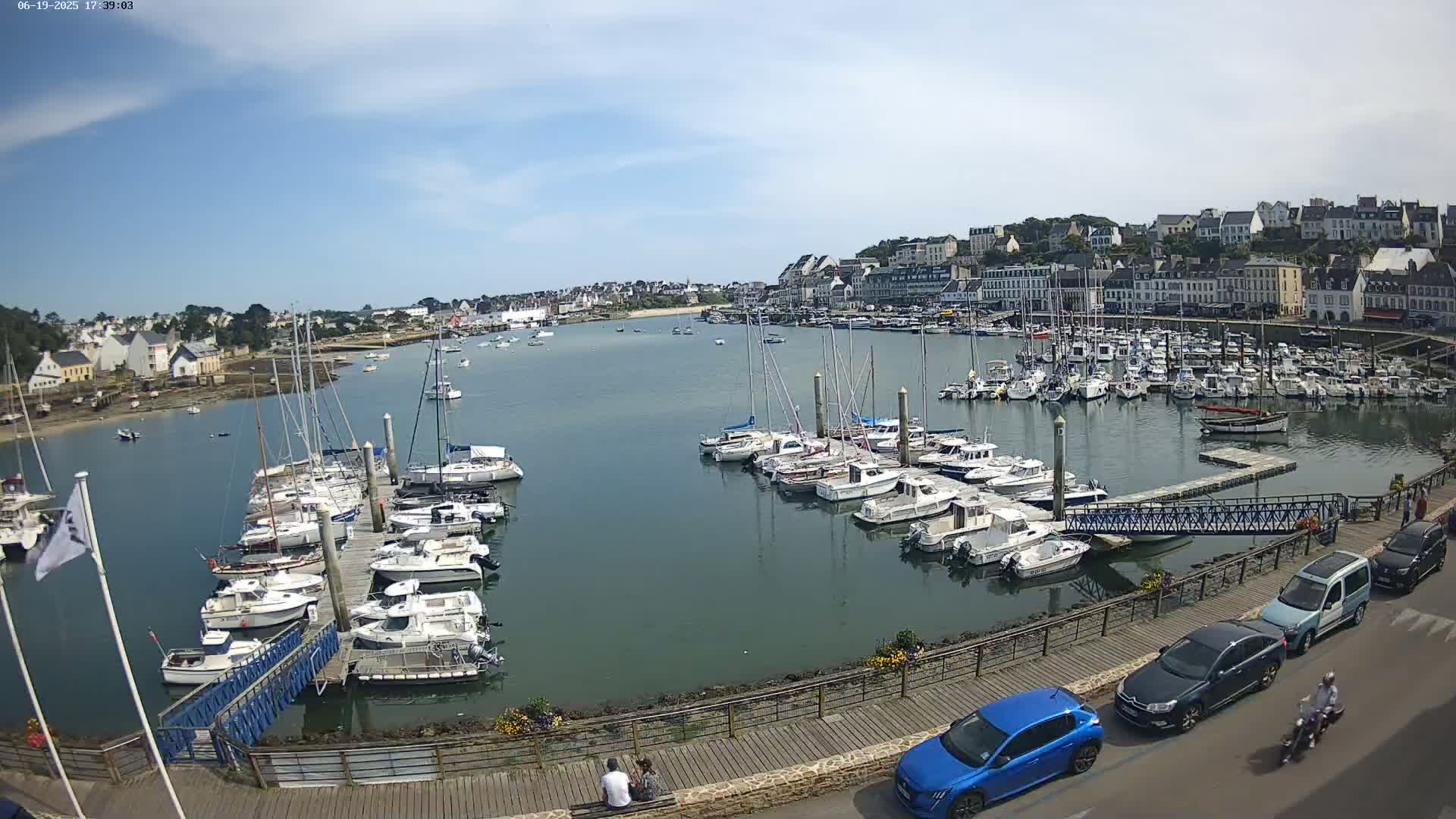 A harbor filled with sailboats and motorboats under a mostly sunny sky, with a town and buildings visible in the background along with a few cars parked along a walkway.