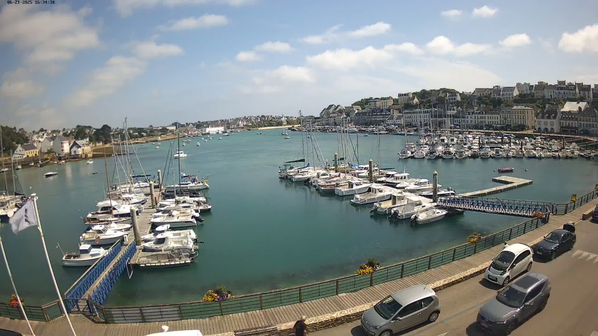 A harbor filled with numerous sailboats and motorboats is nestled next to a town under a partly cloudy sky.