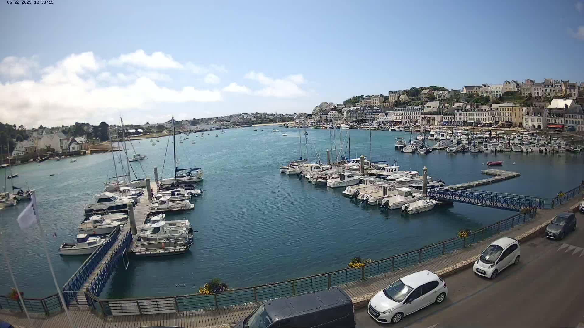 A harbor filled with sailboats and motorboats is situated in front of a town under a mostly sunny sky.