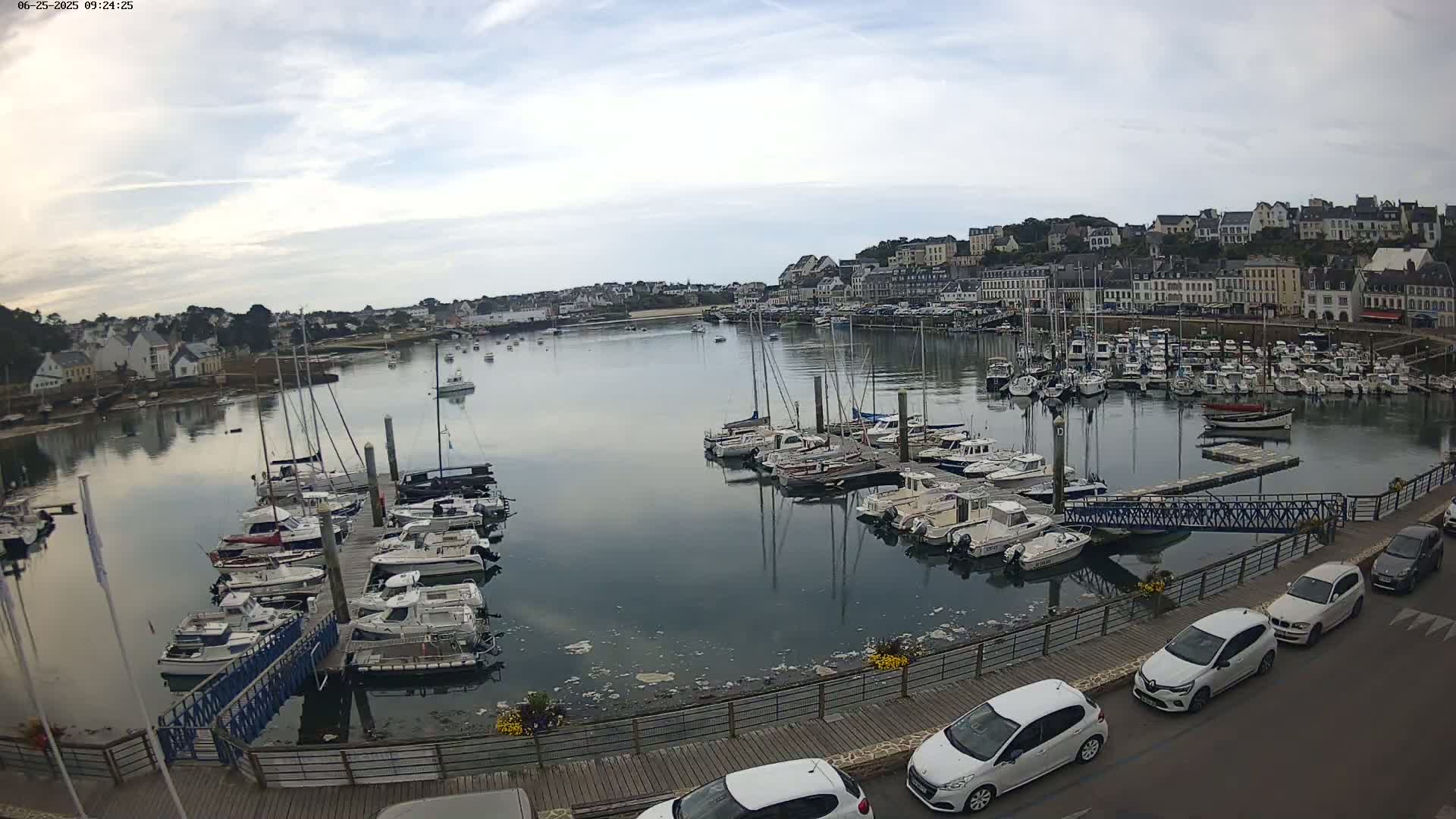A harbor filled with sailboats and motorboats is nestled beside a town under a mostly cloudy sky.