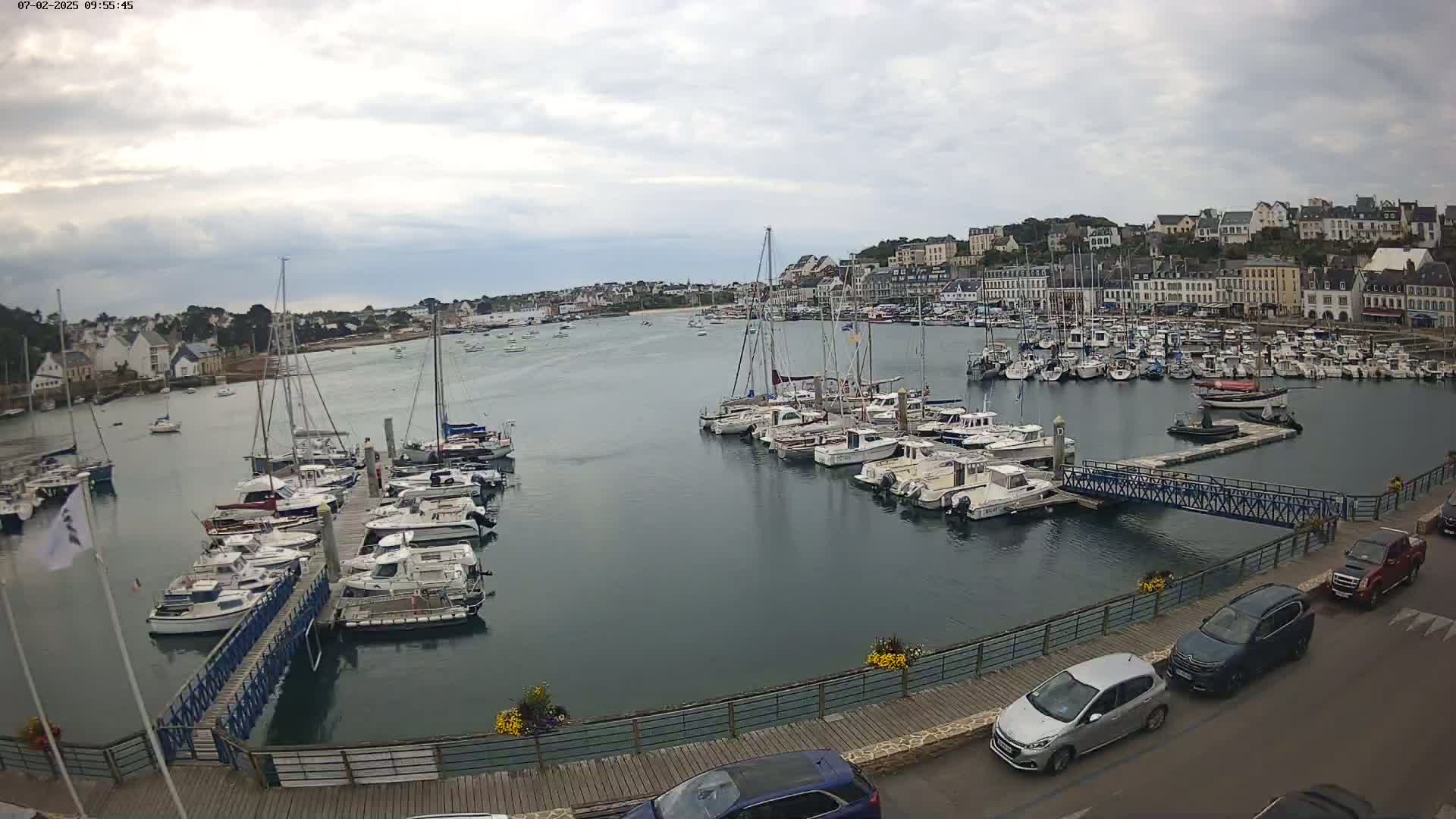 A harbor filled with numerous boats and yachts under an overcast sky, with a town visible in the background and cars parked along a nearby walkway.