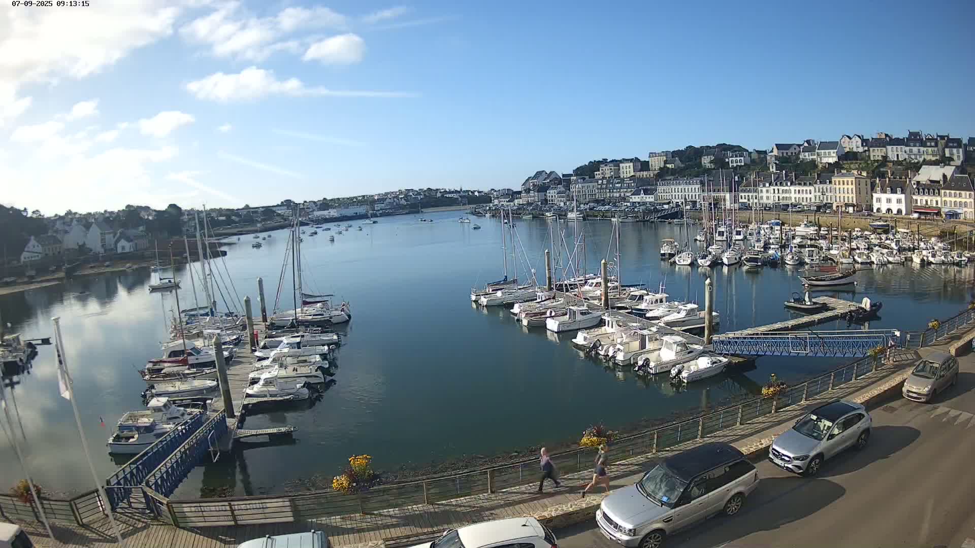 A calm harbor filled with numerous sailboats and motorboats, docked along piers, under a mostly sunny sky, is situated in front of a town with white buildings.
