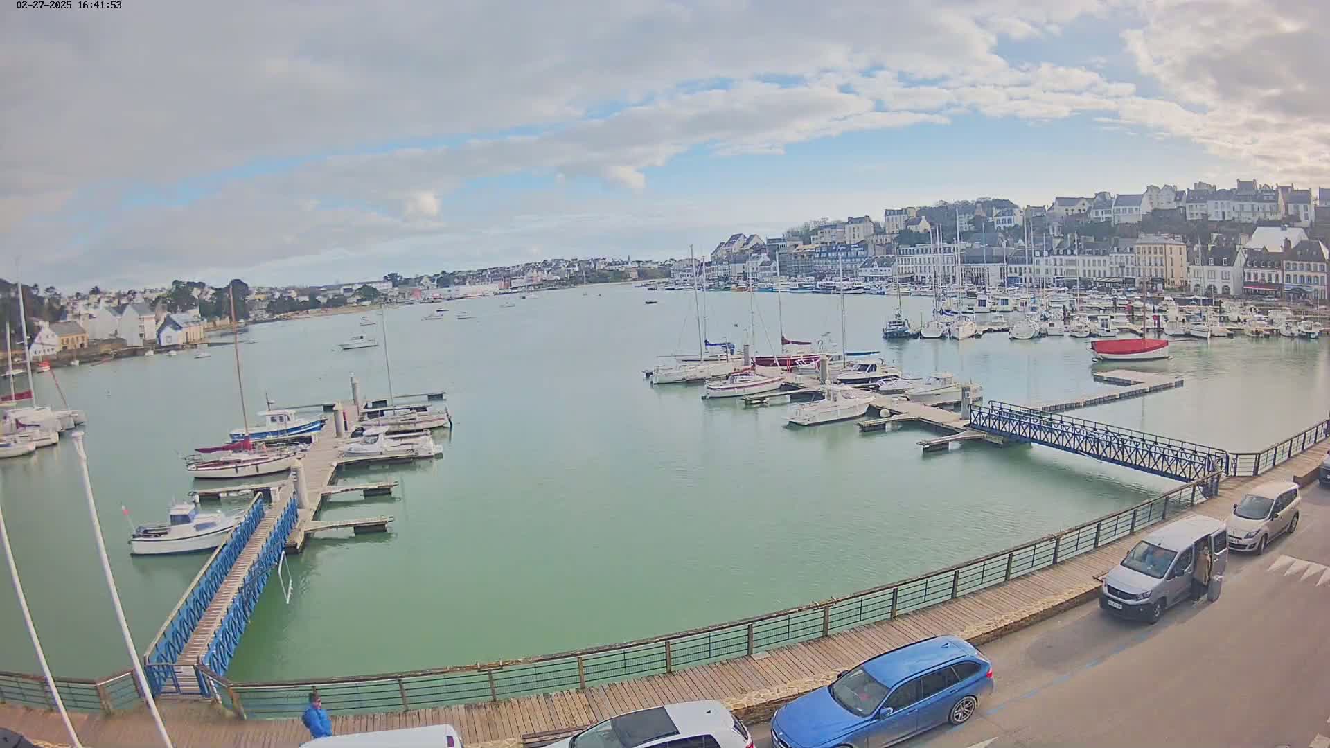 A harbor filled with numerous boats and yachts is situated under a partly cloudy sky, with a town visible in the background.