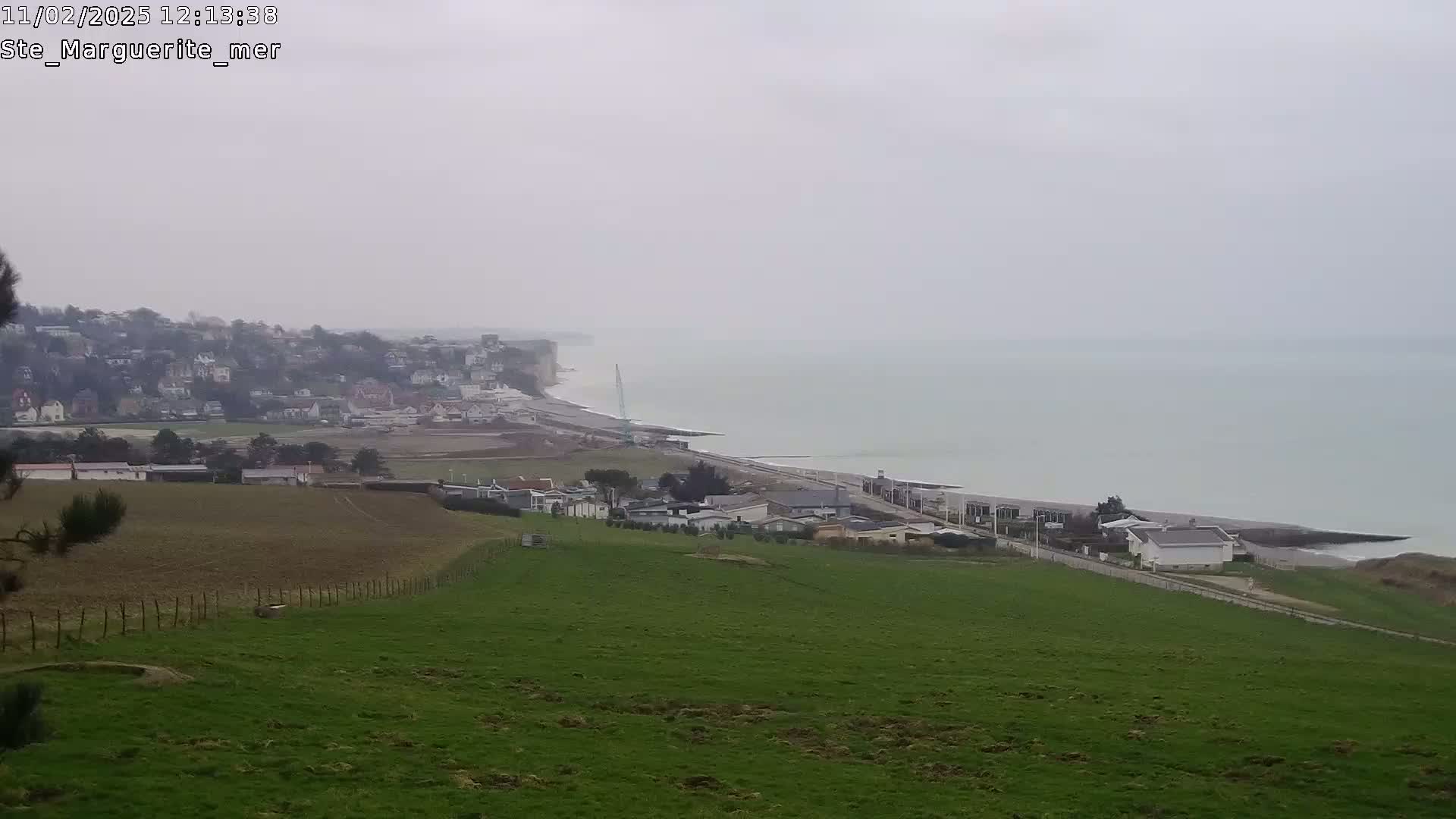 A coastal town sits below a grassy hillside on an overcast day, with a calm sea stretching to the horizon.