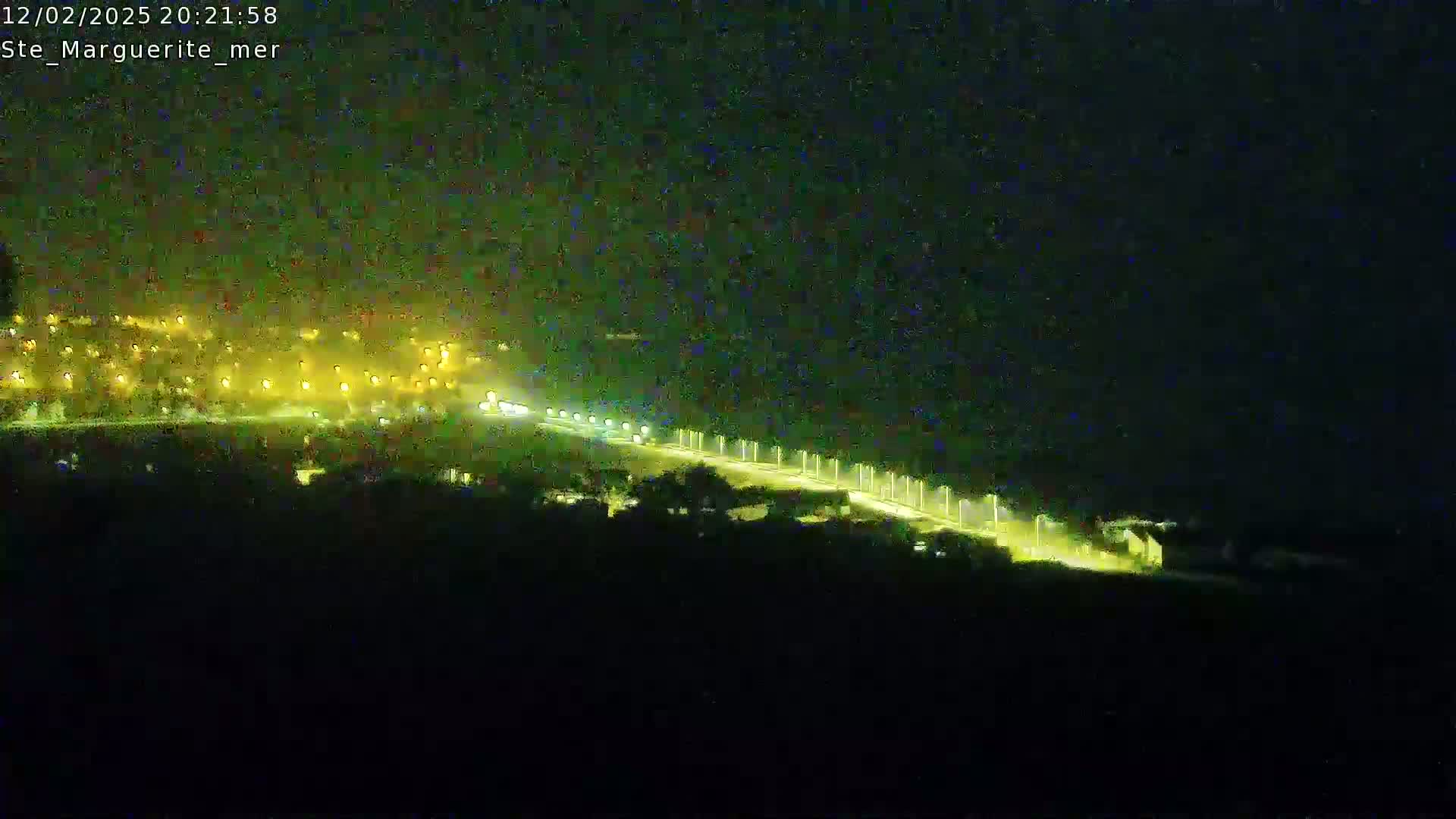 A nighttime, high-angle view shows a brightly lit road curving through a dark landscape, with a town visible in the distance.