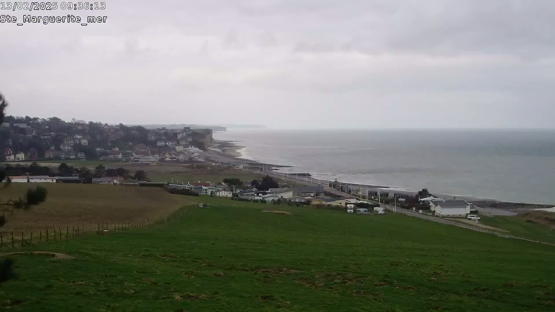 A coastal town sits below a hill overlooking a calm, gray sea on an overcast day.