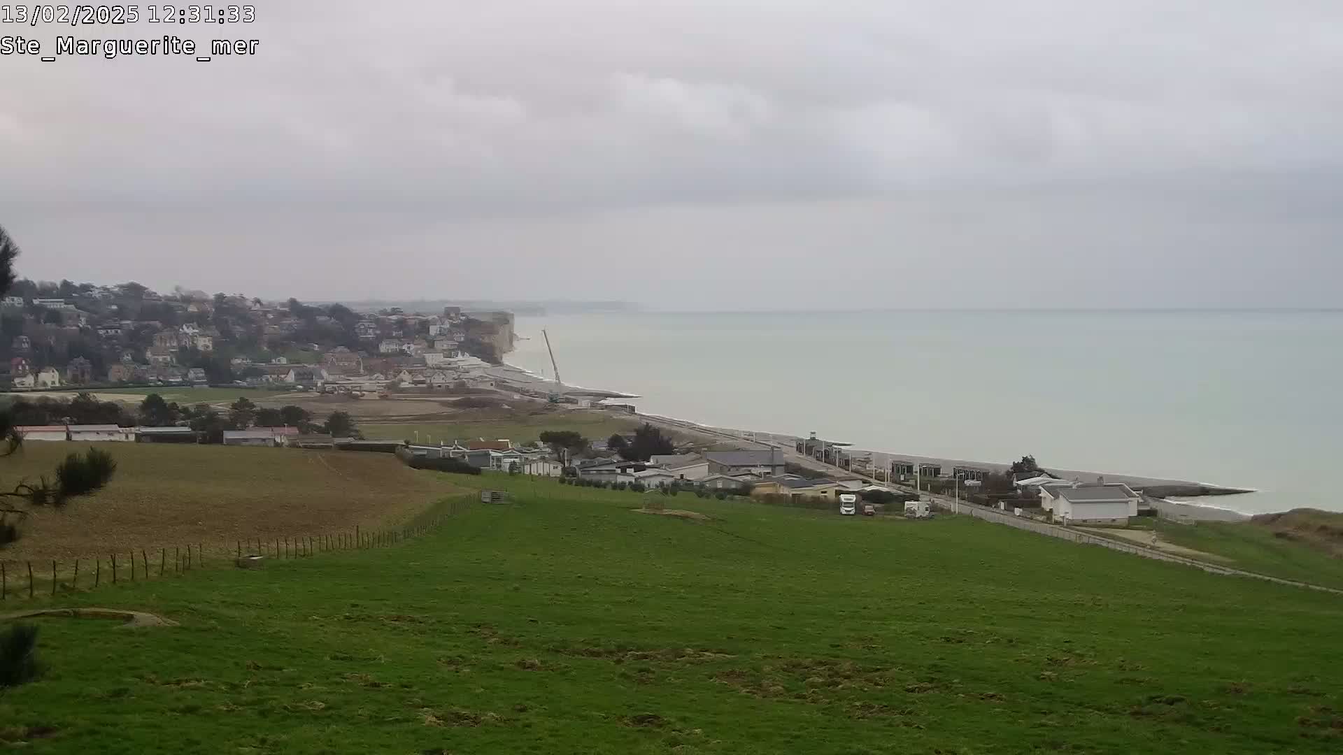 A coastal town sits below a grassy hillside overlooking a calm, overcast sea with white cliffs visible in the mid-ground.