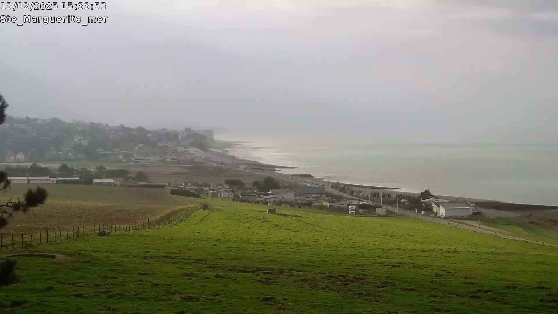 A hazy, overcast day reveals a coastal view of a town nestled beside a calm sea, with rolling green fields stretching towards the viewer.