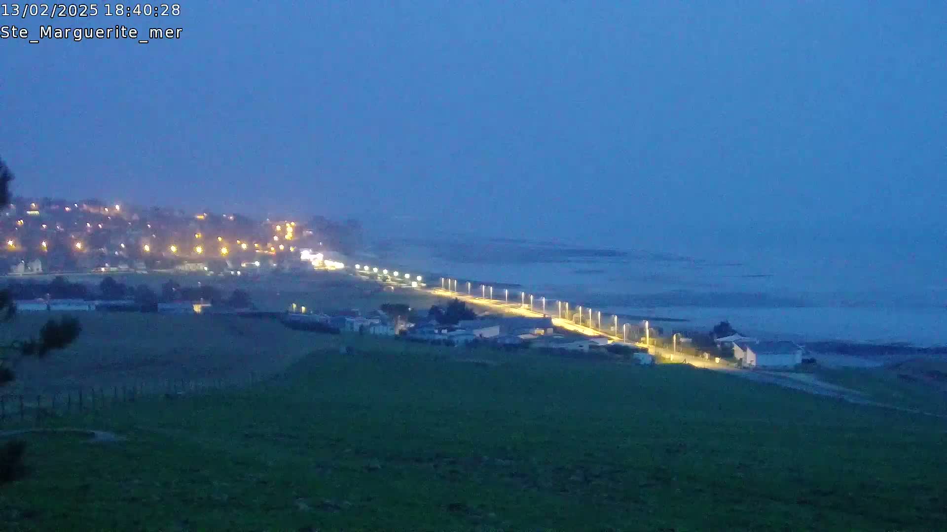 A coastal town at twilight is illuminated by streetlights along a road next to the ocean under a dusky sky.