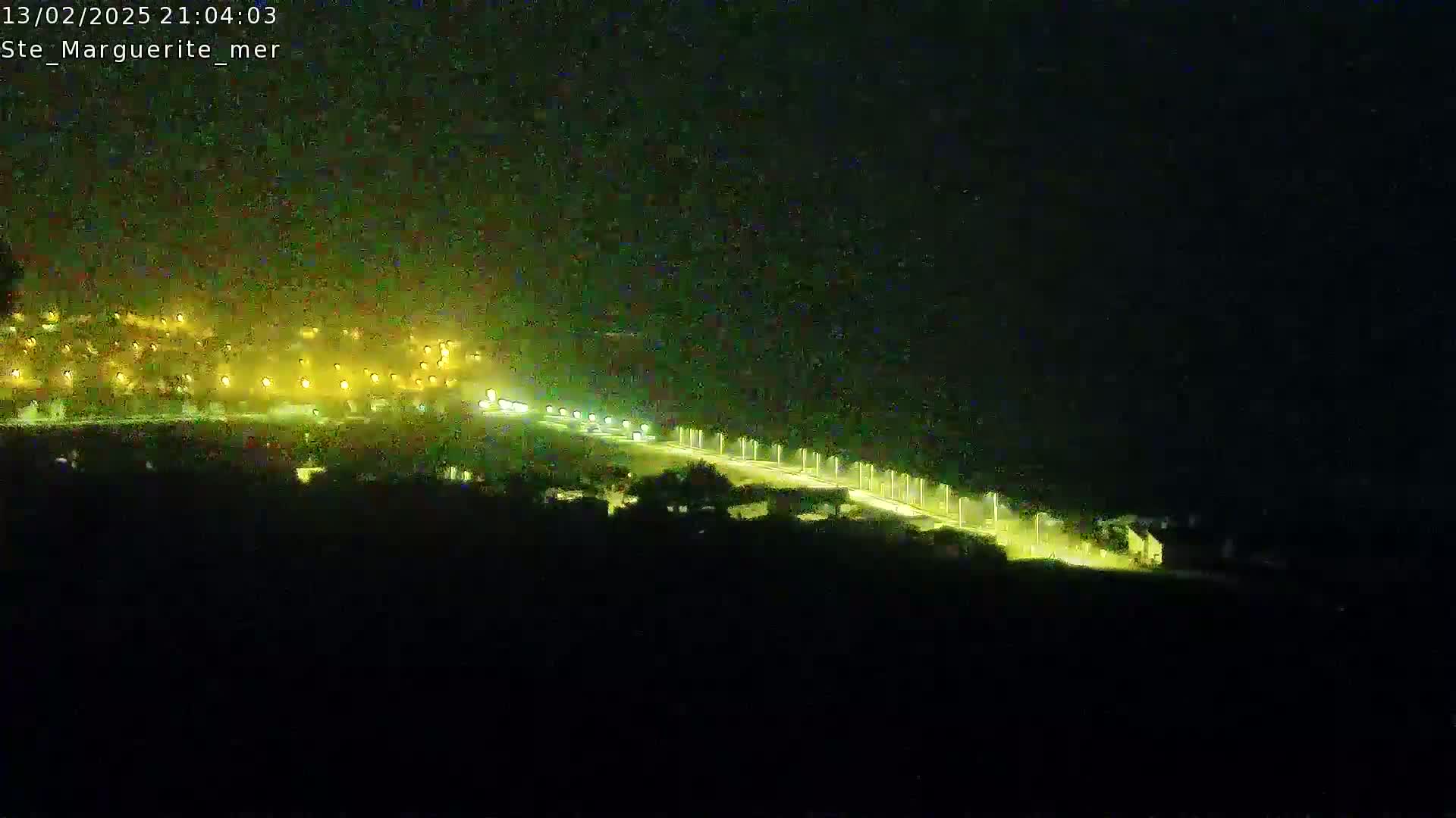 A nighttime, high-angle view shows a brightly lit road curving through a dark landscape, with a town's lights visible in the distance.