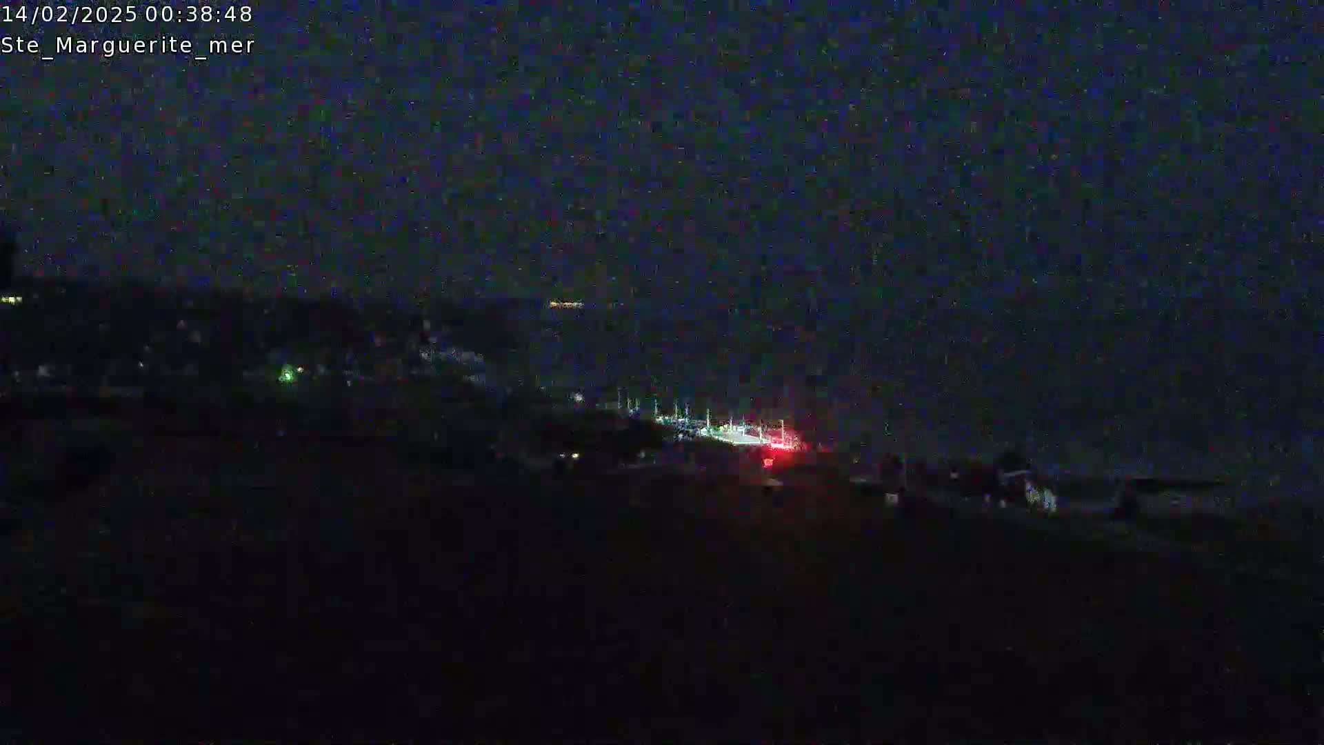A dark, nighttime scene shows a road with a few lights visible, leading down towards a dark body of water under a clear, starless sky.