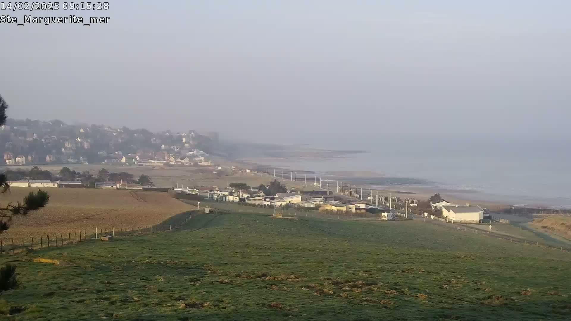 A hazy coastal scene shows a village nestled between a grassy field and a calm sea under a pale sky.