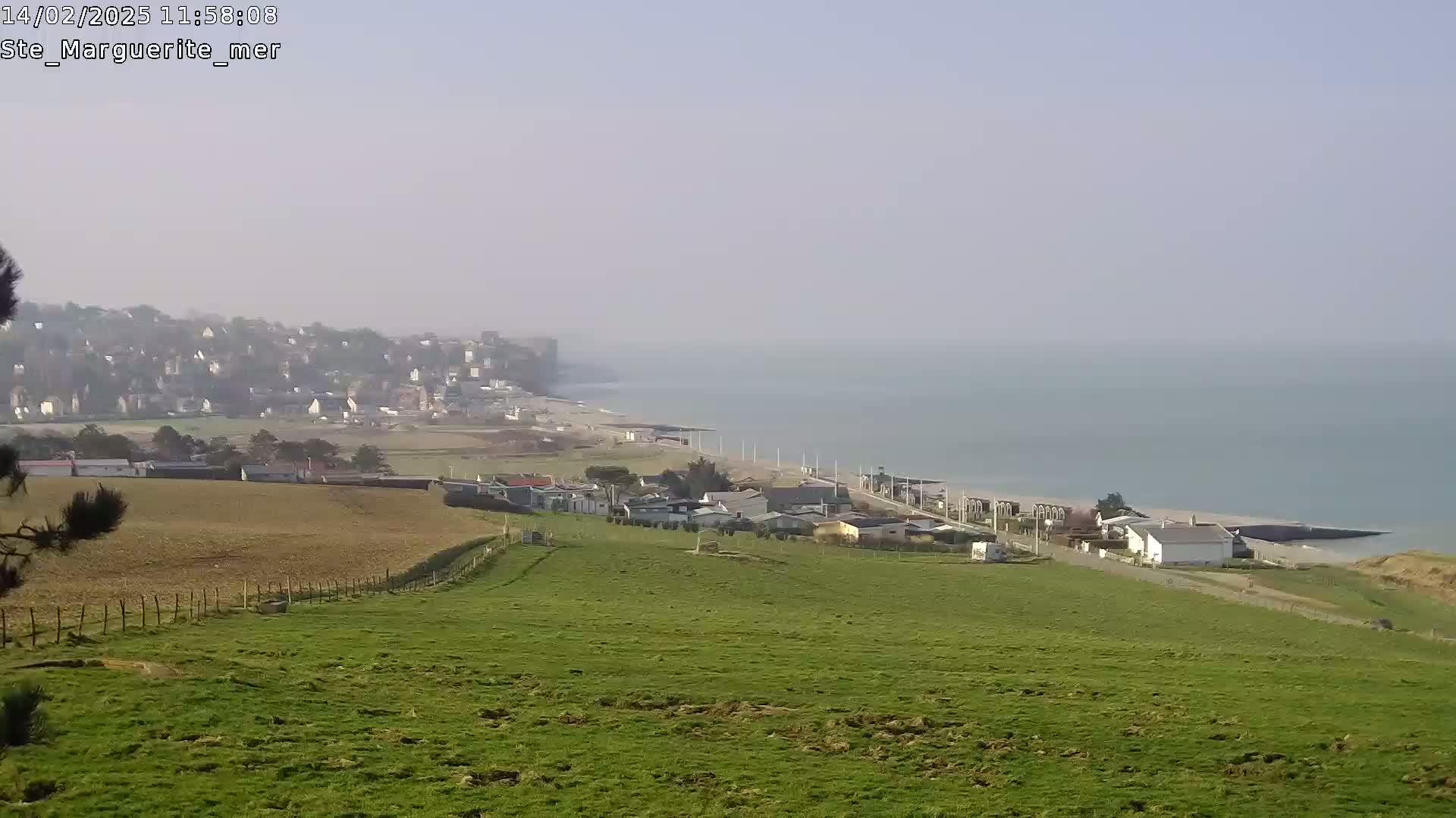 A hazy, coastal scene shows a green hillside overlooking a town, beach, and calm sea.