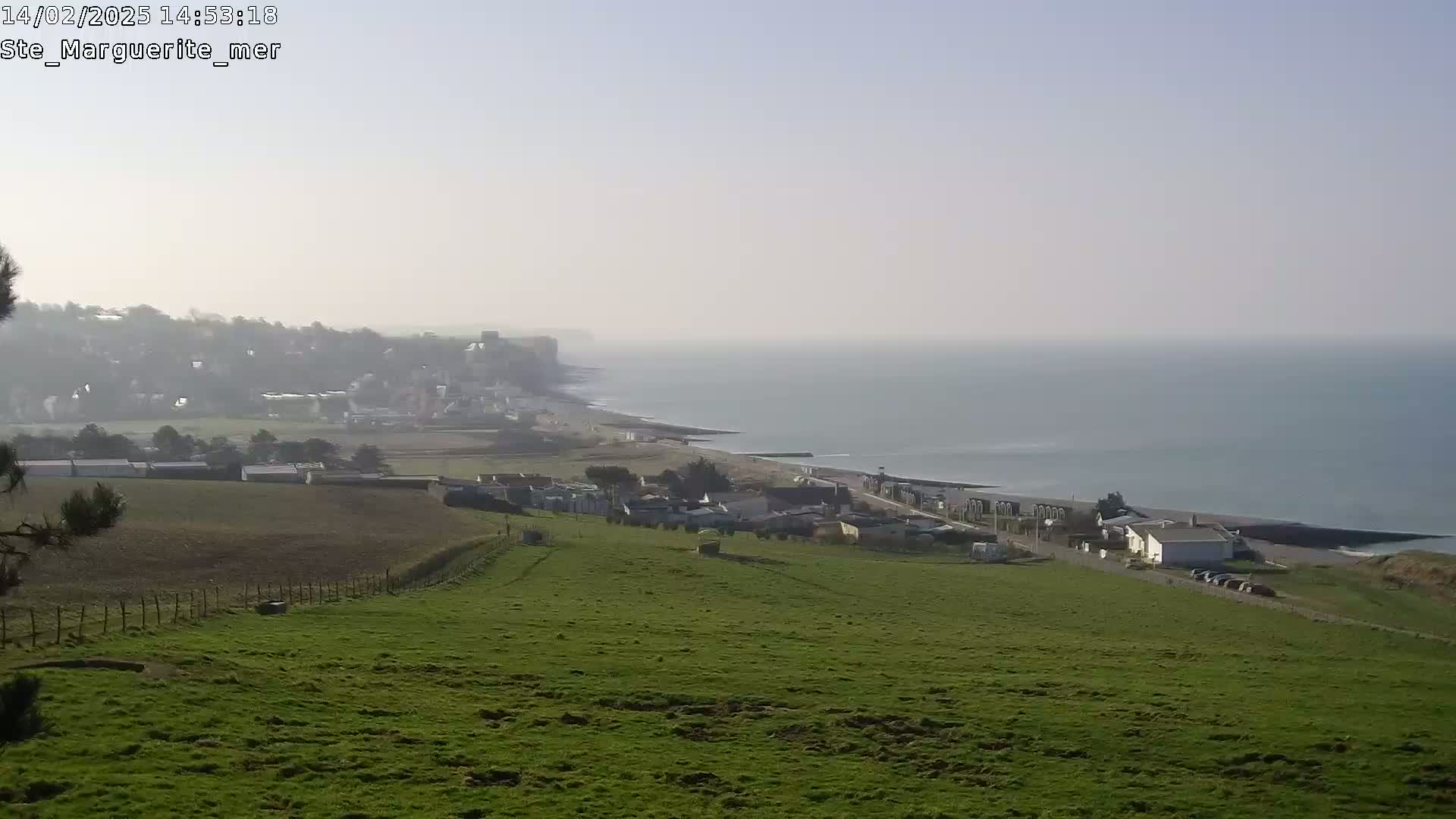 A hazy view shows a green hillside overlooking a coastal town and a calm sea stretching to the horizon.