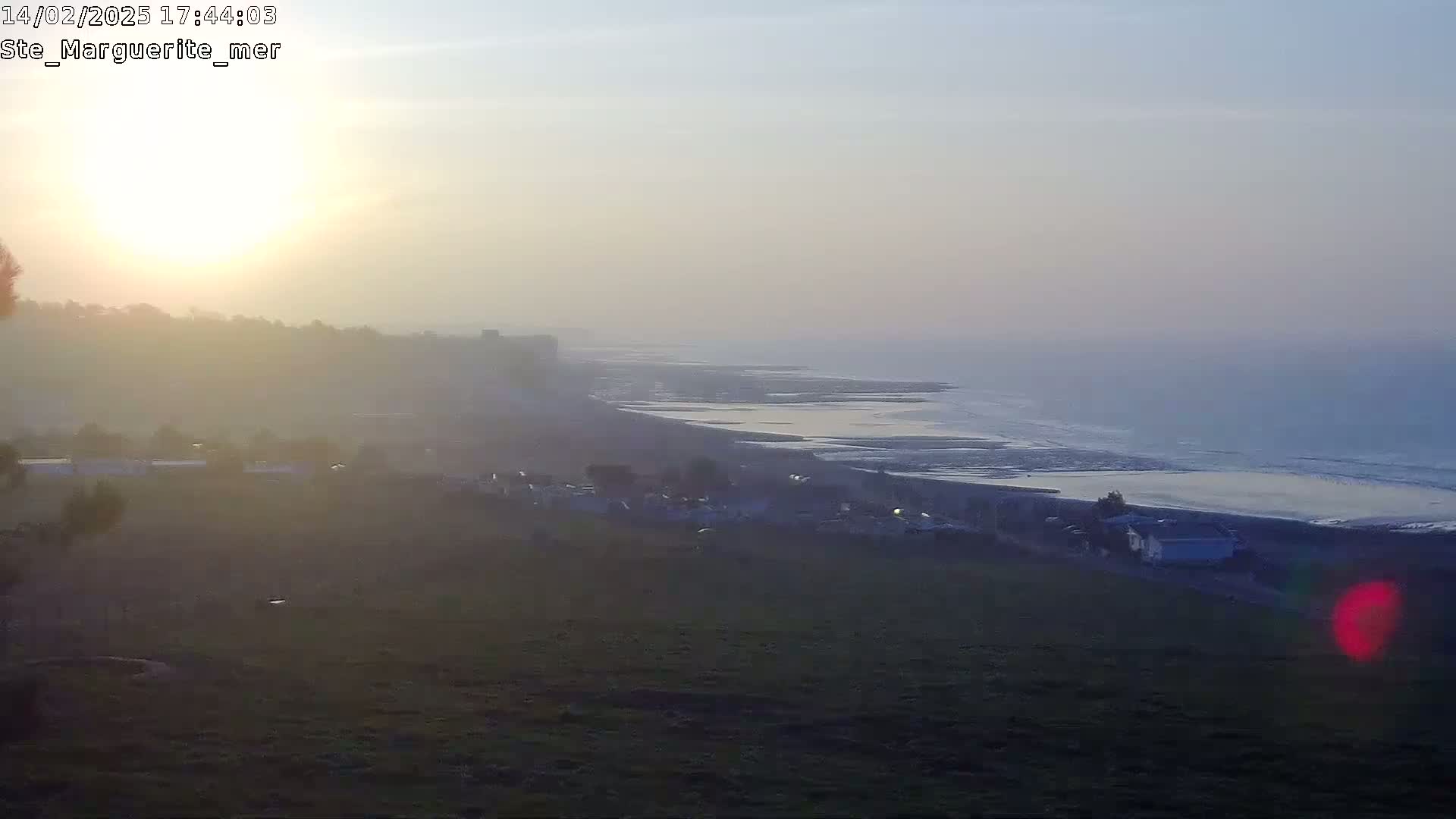 A hazy sunset casts a golden glow over a coastal landscape, showing a beach, ocean, and distant hills.