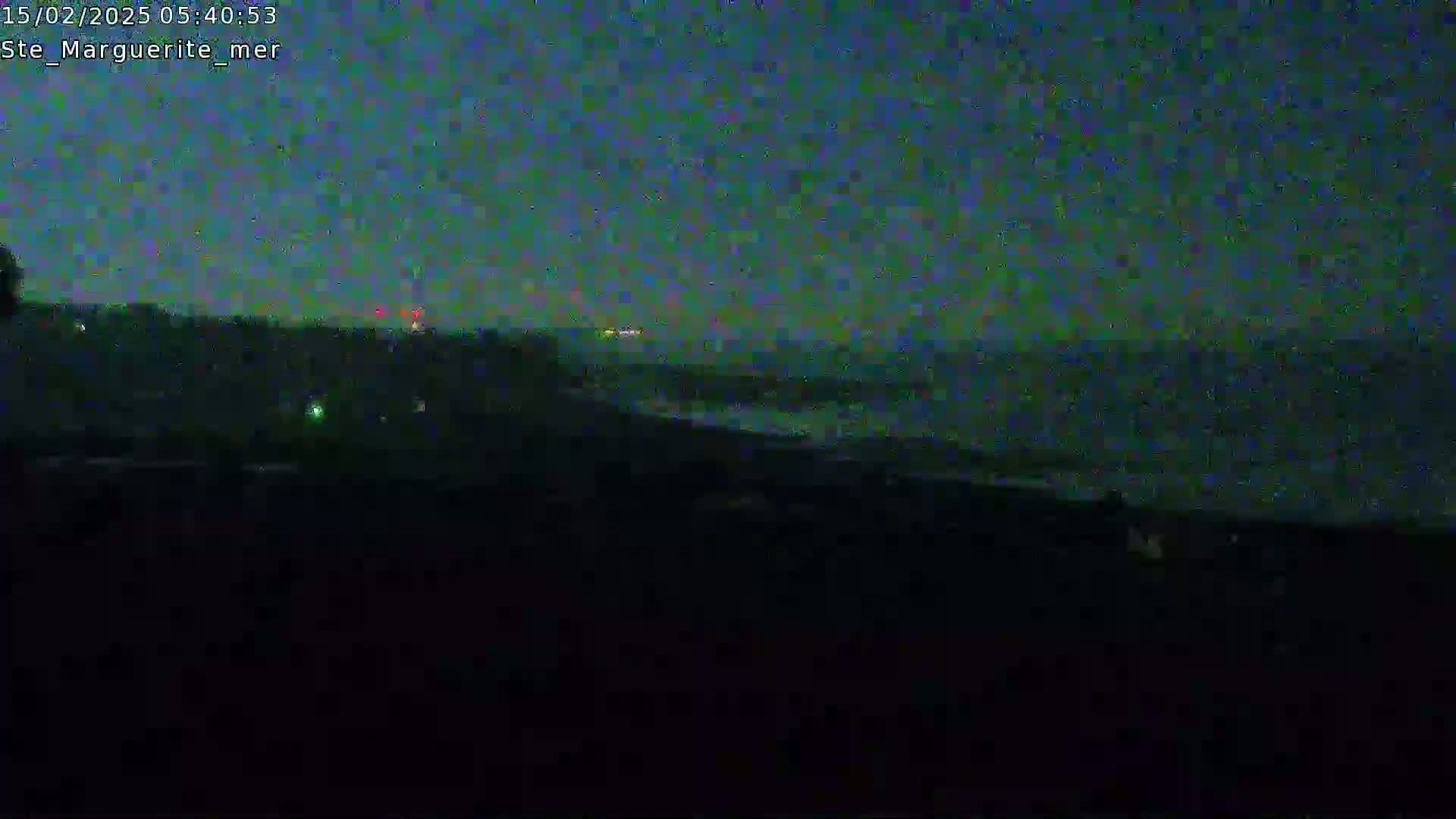 A dark, nighttime view of a shoreline with a body of water showing faint waves under a mostly clear, dark sky.