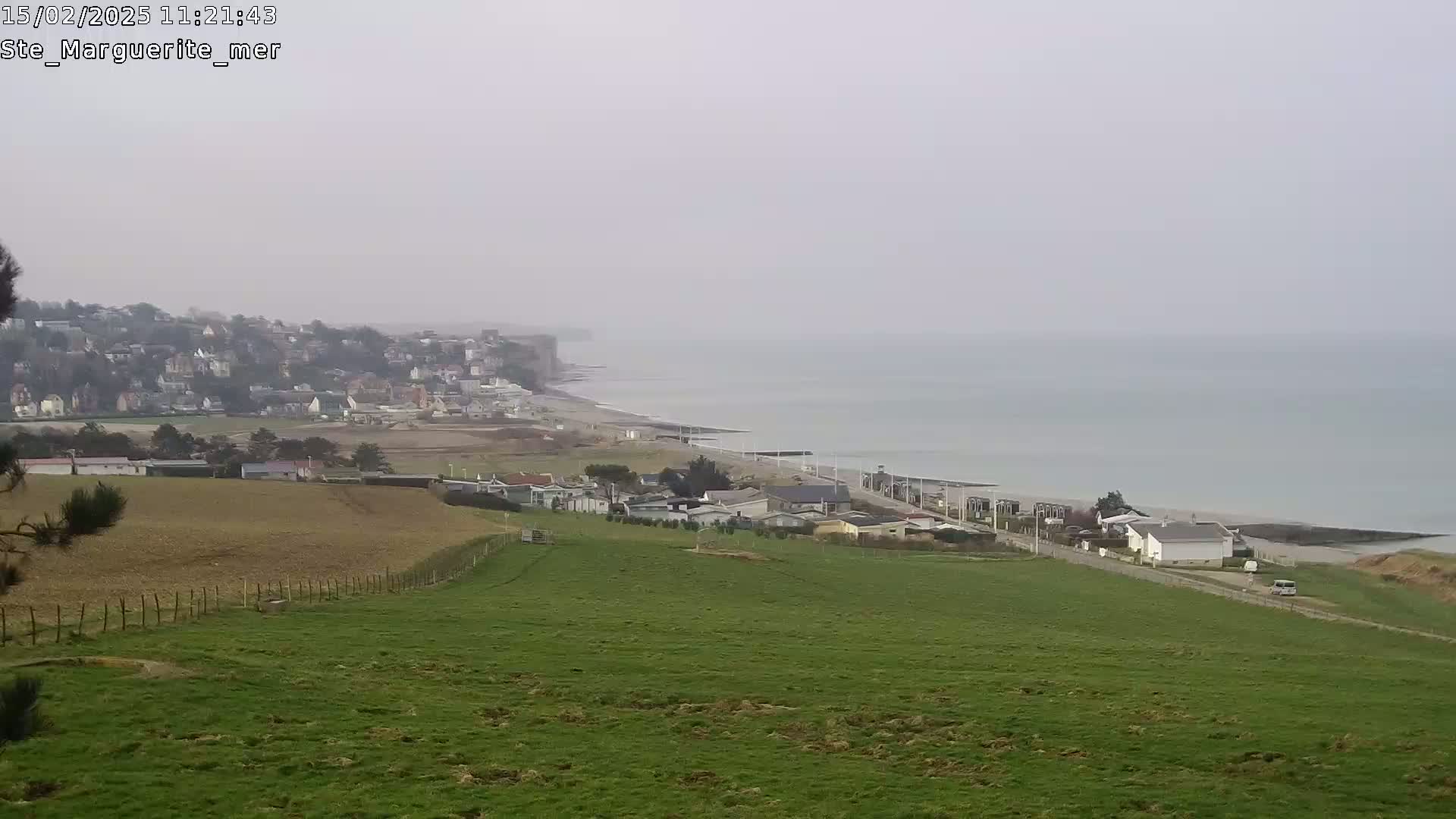 A hazy coastal view shows a green pasture overlooking a town nestled below white cliffs and a calm ocean.