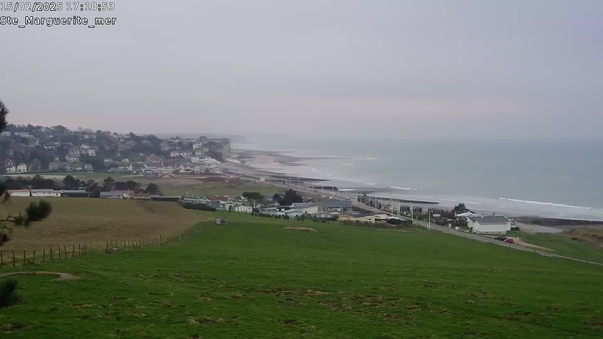 A coastal town and beach are visible from a grassy hillside under an overcast sky.