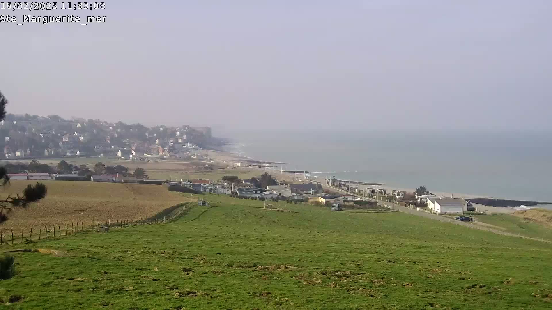 A hazy, coastal view shows a green hillside overlooking a town, a beach, and the ocean.