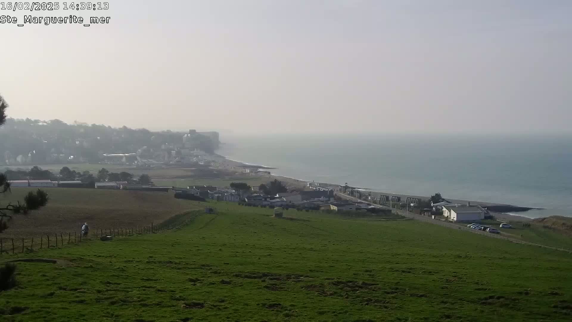 A hazy coastal view shows a green hillside overlooking a village, a beach, and a calm sea under a light, overcast sky.