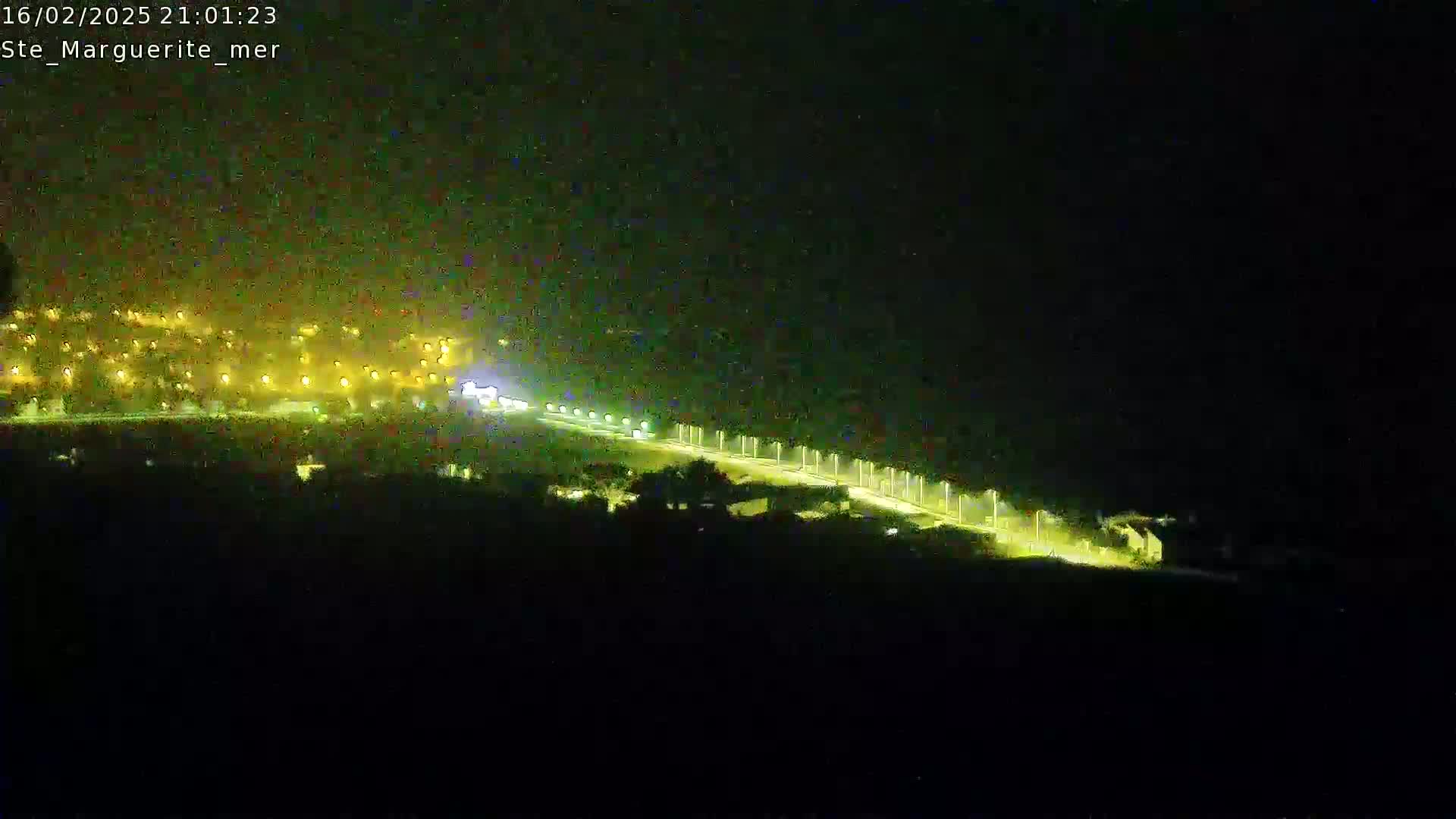A nighttime, high-angle view shows a dimly lit road winding through a dark landscape toward a village with scattered lights.