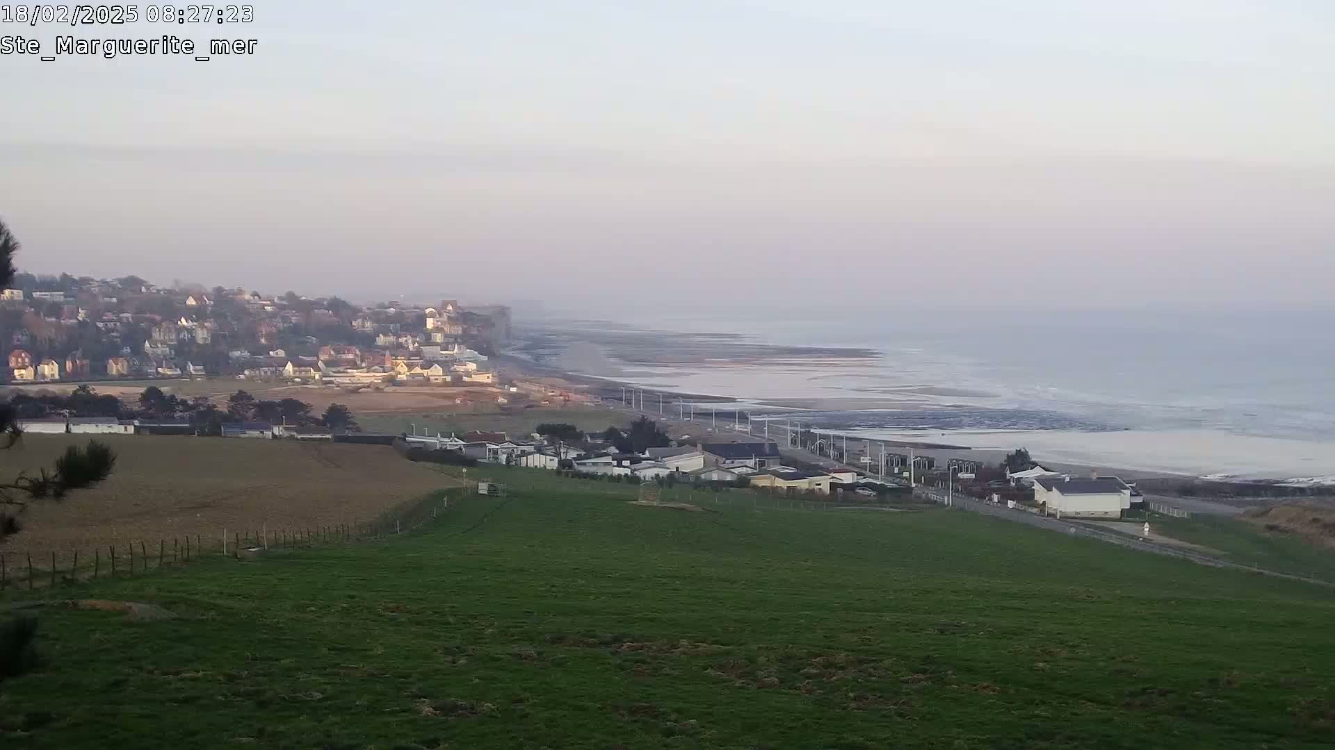 A coastal town with houses situated on a cliff overlooking a calm, light-grey ocean under a hazy sky.