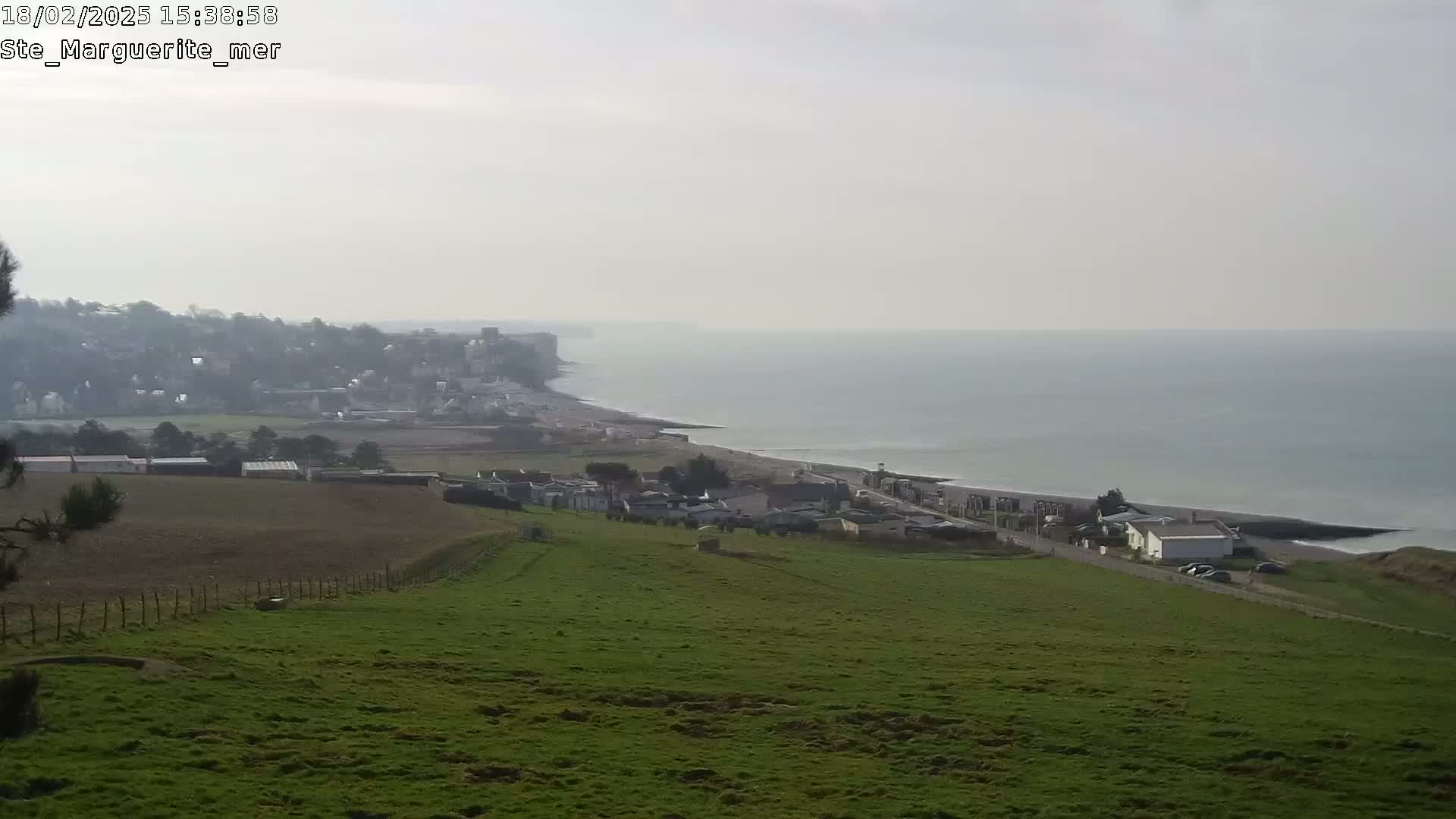 A hazy coastal view shows a village nestled on a green hillside overlooking a calm sea and a distant town along a curving shoreline.