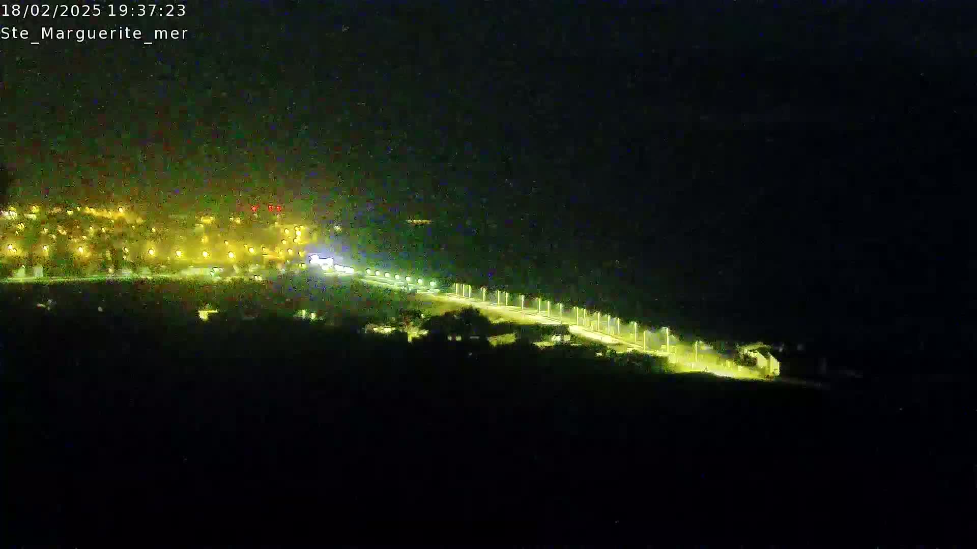 A nighttime, high-angle view shows a brightly lit road curving through a dark landscape, with a town's lights visible in the distance.