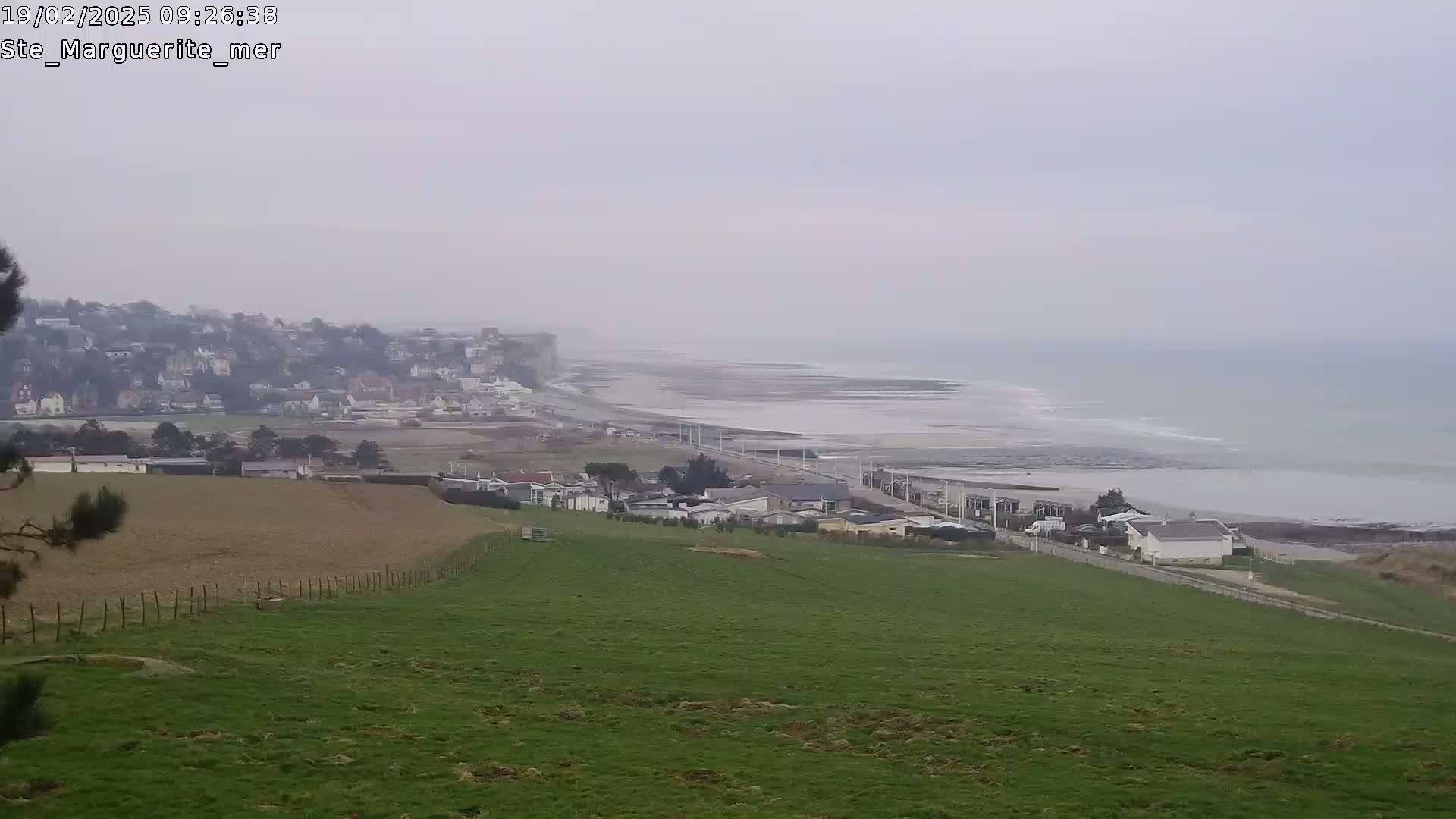 A hazy coastal scene shows a town nestled against a cliff overlooking a beach and ocean with gentle waves under an overcast sky.