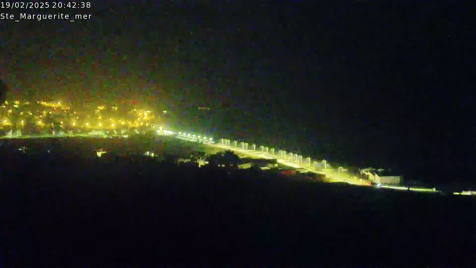 A nighttime, long-range view of a lit roadway, with a town’s lights visible in the distance.