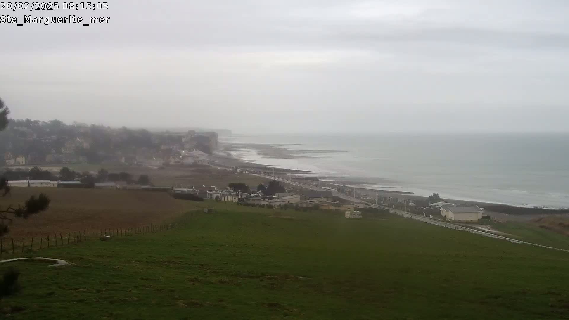 A coastal scene on an overcast day shows a stretch of gray ocean meeting a beach and a road that winds past houses and farmland.