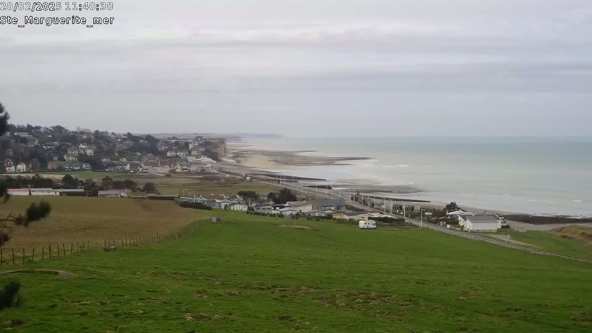 A coastal town sits below a verdant hillside overlooking a calm, gray sea on an overcast day.
