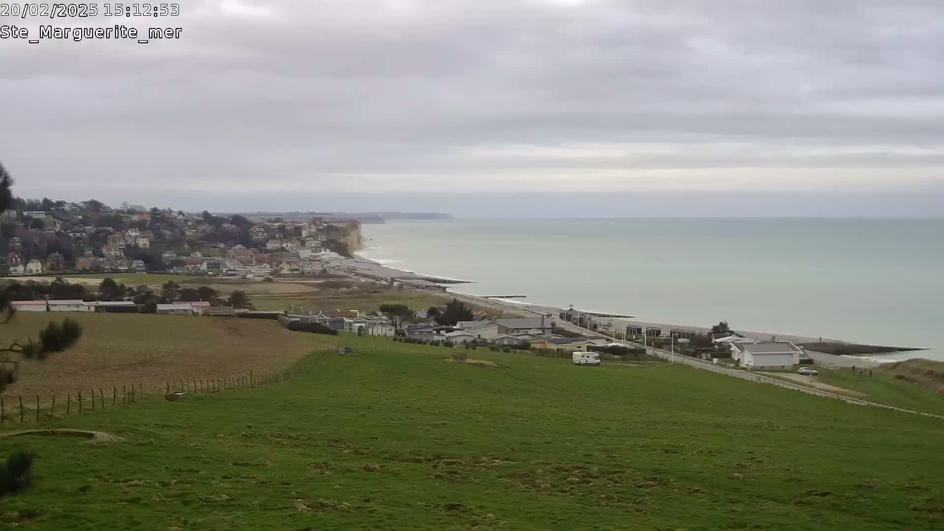 A coastal town nestled below white cliffs overlooks a calm, light-green sea under an overcast sky.