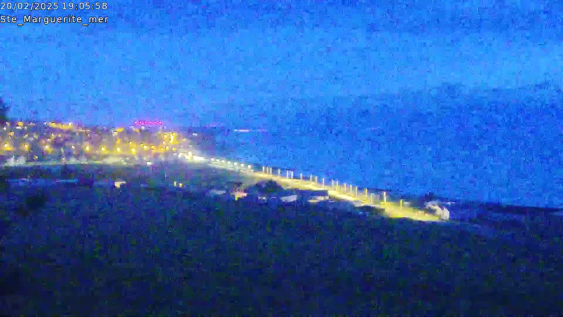 A nighttime coastal scene shows a dark, land-based area with lights, a dark body of water, and a dimly lit, paved path extending from the land toward the water under a dark blue, twilight sky.
