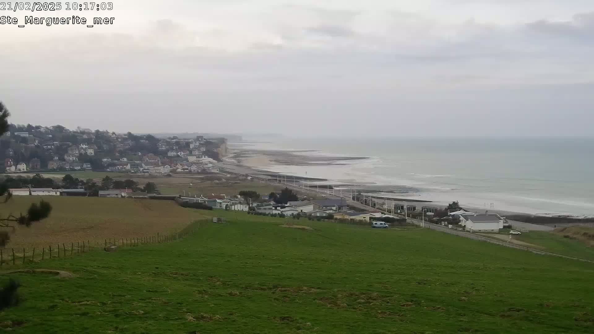 A coastal town sits atop a cliff overlooking a gray, overcast ocean with gentle waves lapping at a sandy beach.