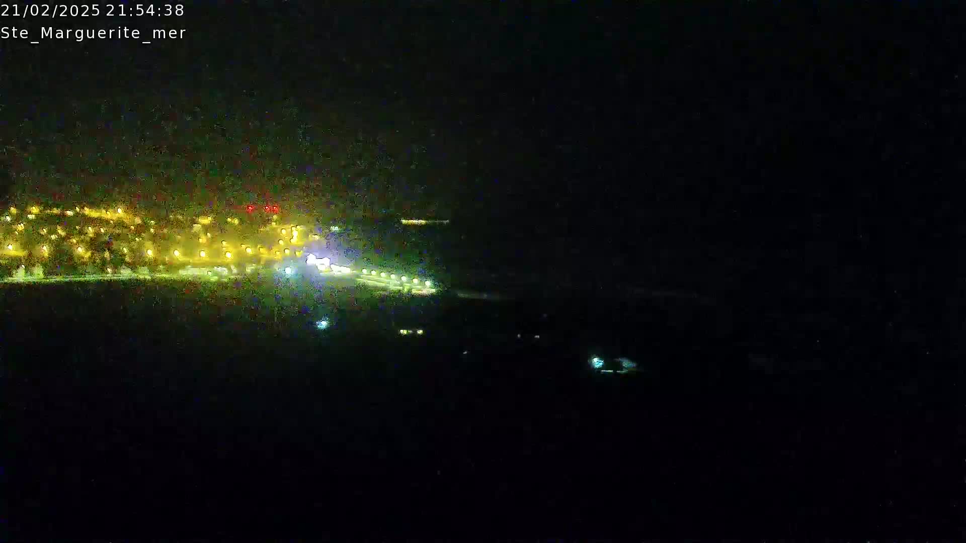 A nighttime view of a coastal town with its lights visible, a dark body of water in the foreground, under a dark sky.